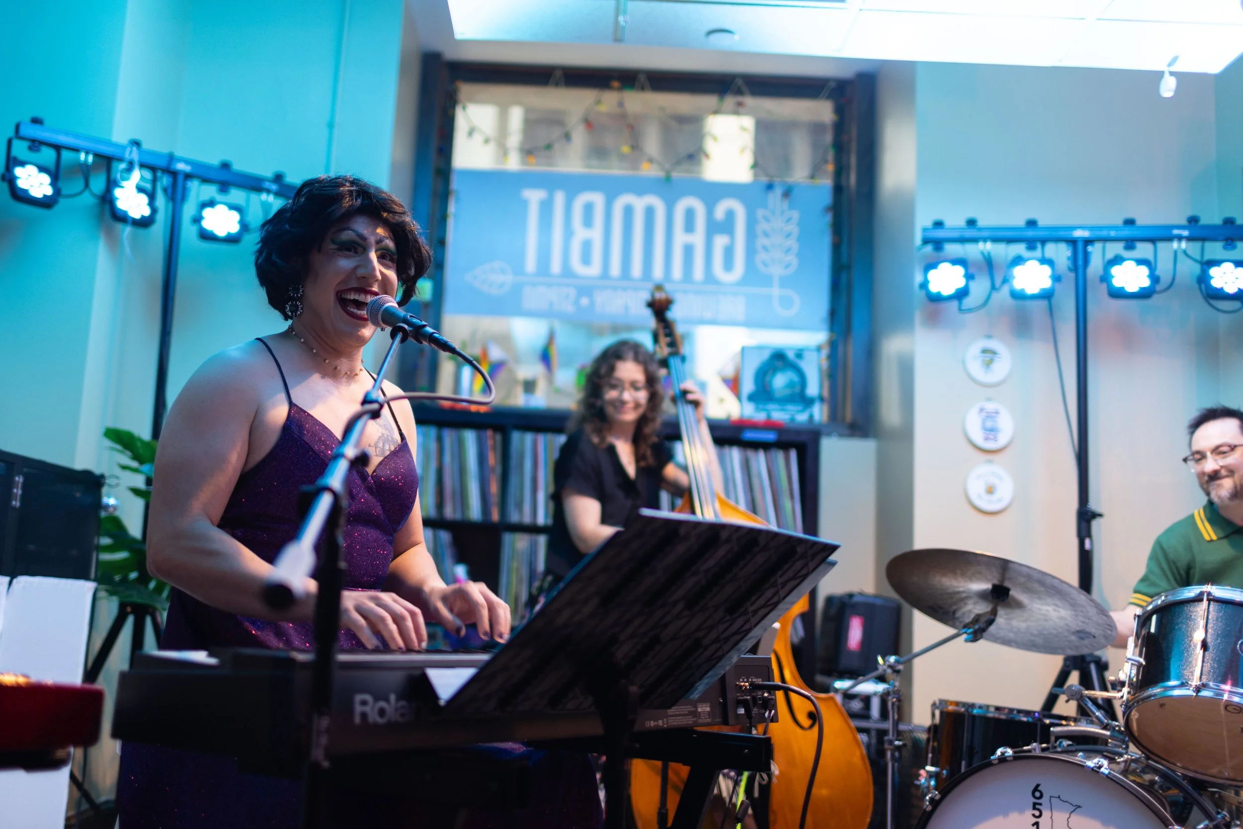 A group of musicians performing indoors. A woman in a purple dress with short dark hair is singing into a microphone, playing a keyboard. Behind her, a woman with curly hair is playing a double bass, and a man with glasses is playing drums. There is a large banner in the background with the word 'CAMPB' visible, and bright blue stage lights are illuminating the scene.