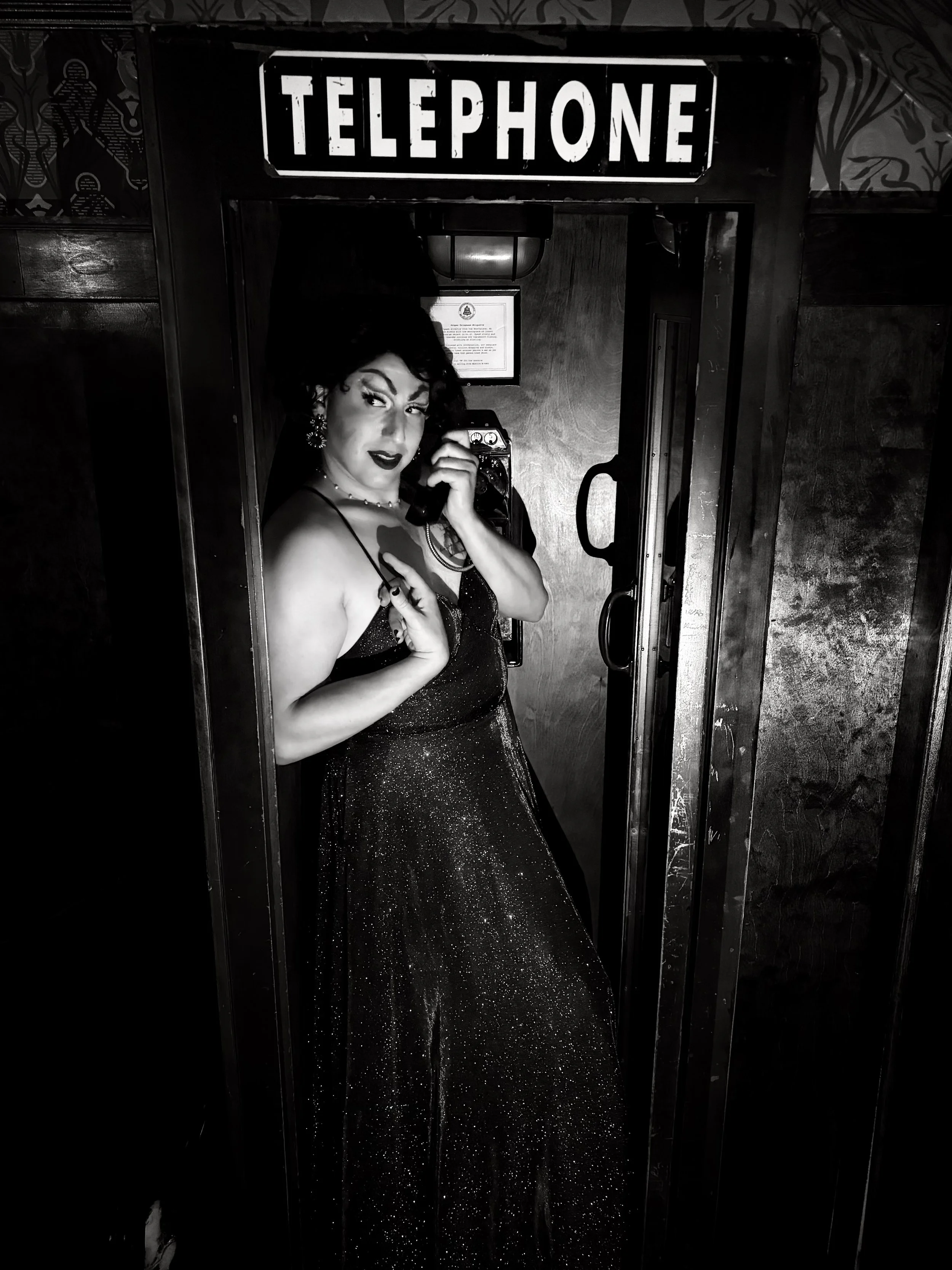 Black and white photo of Blanche with styled hair and makeup, standing in front of a telephone booth, holding a vintage mobile phone to her ear, in a glamorous, 1950s-inspired outfit and jewelry.