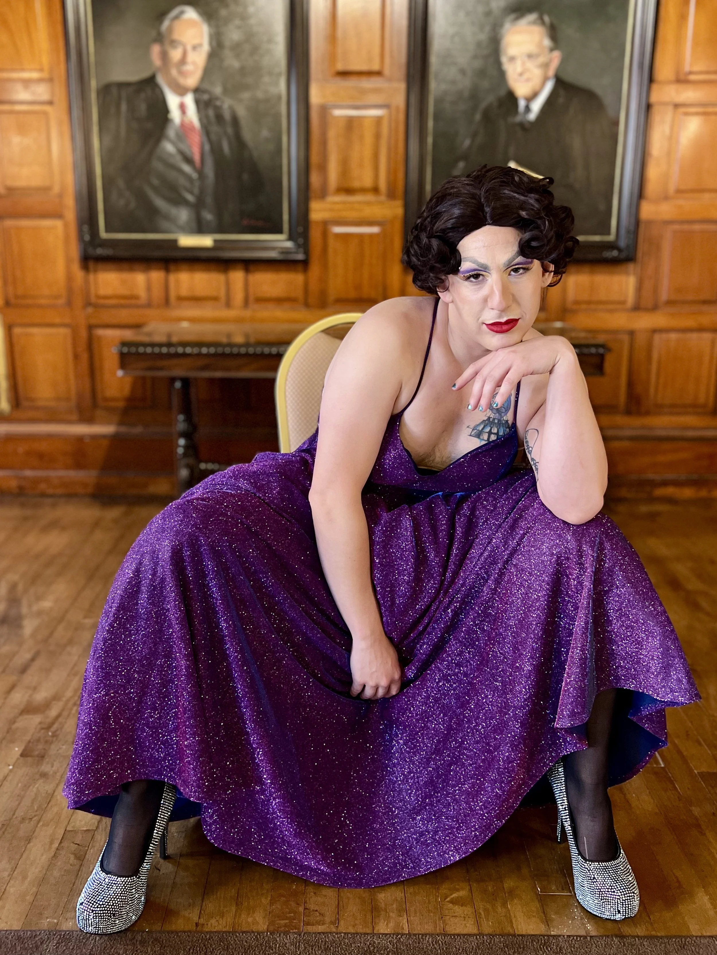 Person with short black curly hair, makeup, and a red lipstick, wearing a shiny purple dress, sitting on a chair with legs apart, inside a room with wooden walls and portraits of two men hanging on the wall behind.