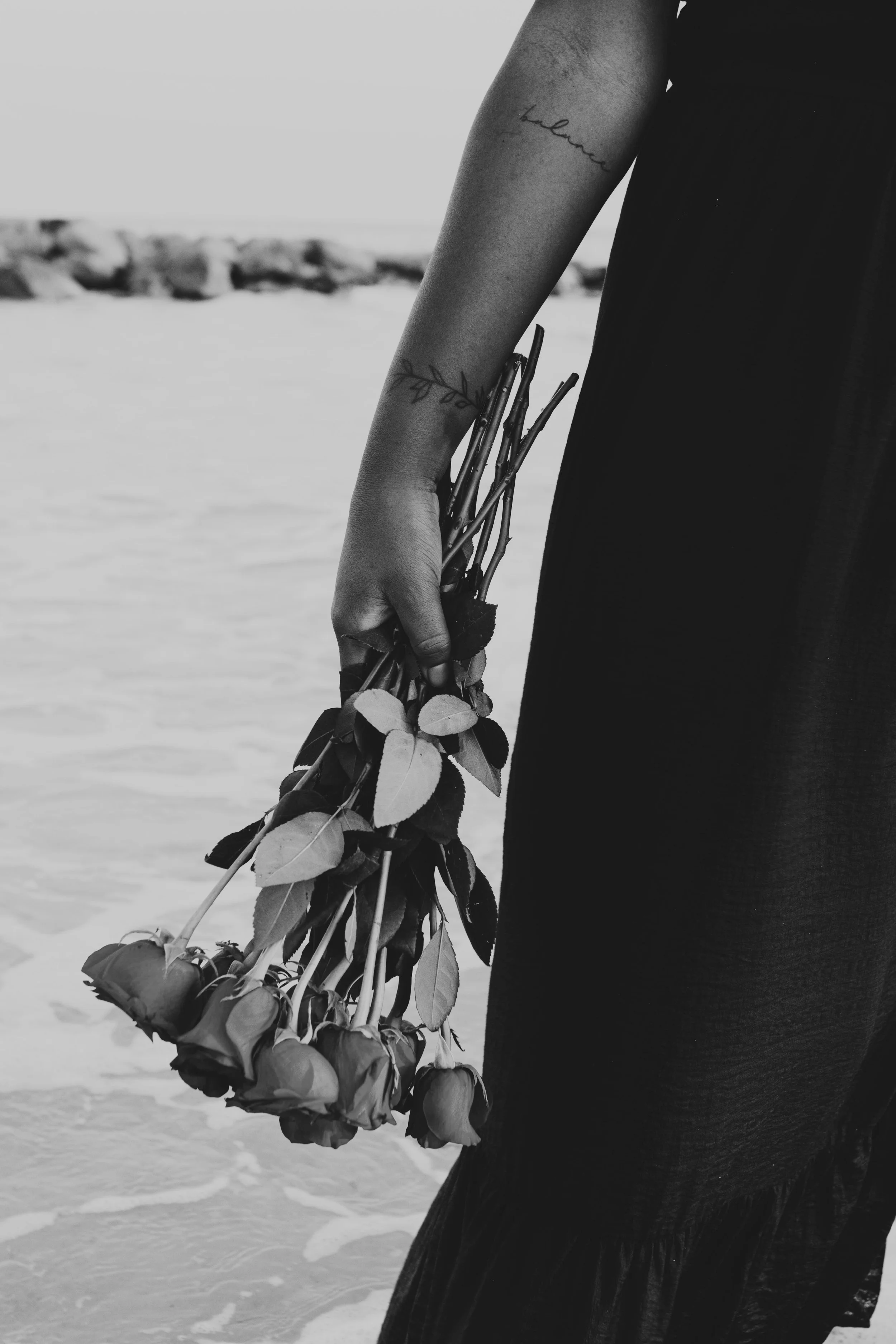 A person holding a bouquet of roses, wearing a dark dress, standing on a beach with rocks in the background. The person has a tattoo on their forearm that reads 'hello', and another tattoo of a leafy branch on the same arm.