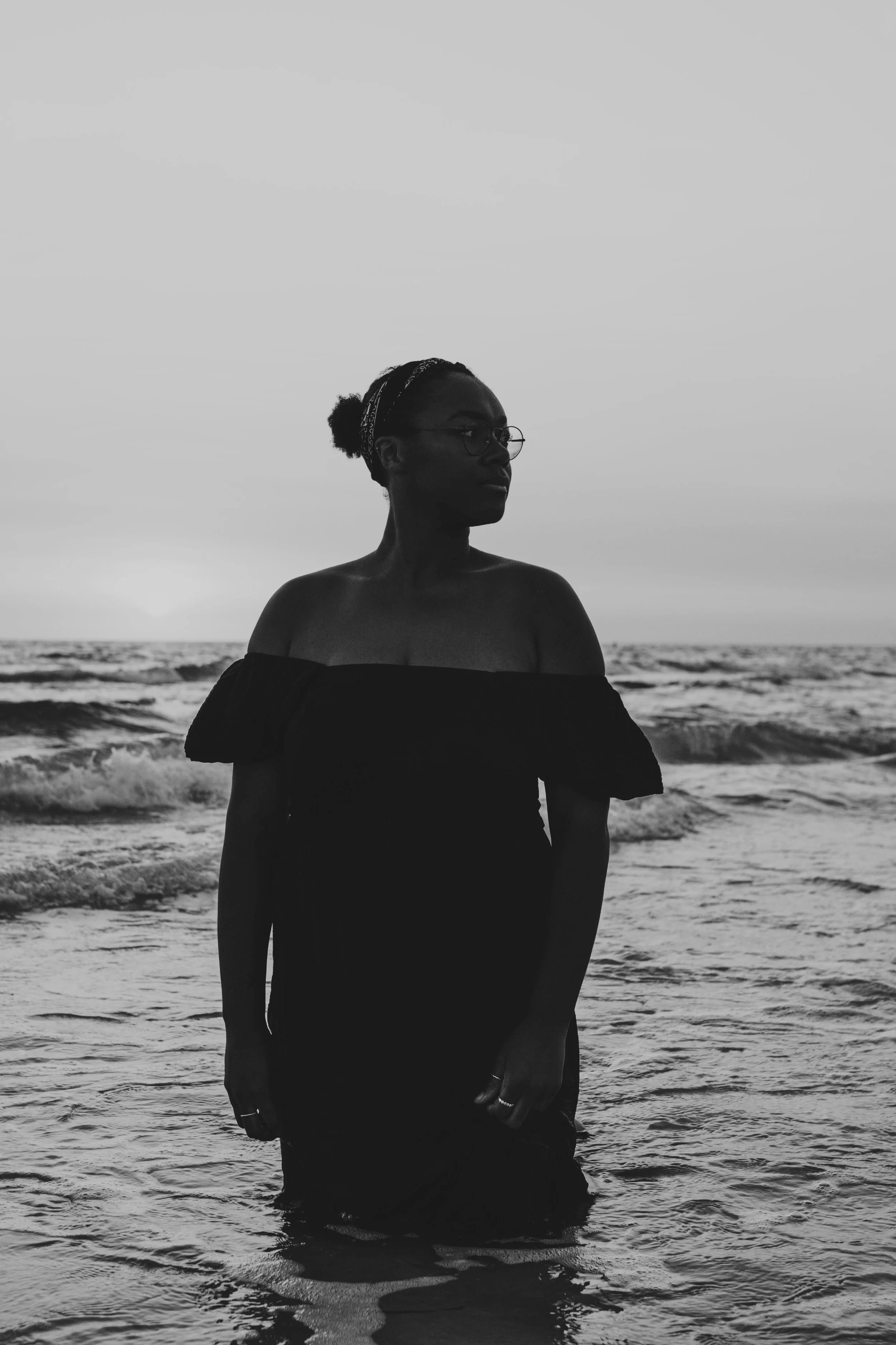 A woman standing in the ocean at sunset, looking to the right, wearing glasses and an off-the-shoulder dress, with waves around her legs.