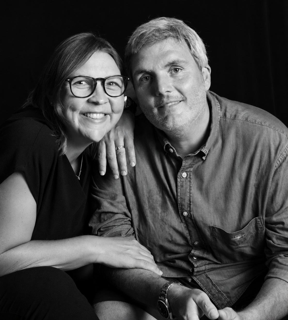 A black-and-white photo of a smiling woman with glasses and shoulder-length hair and a man with short hair and a beard, both posing closely together against a dark background.