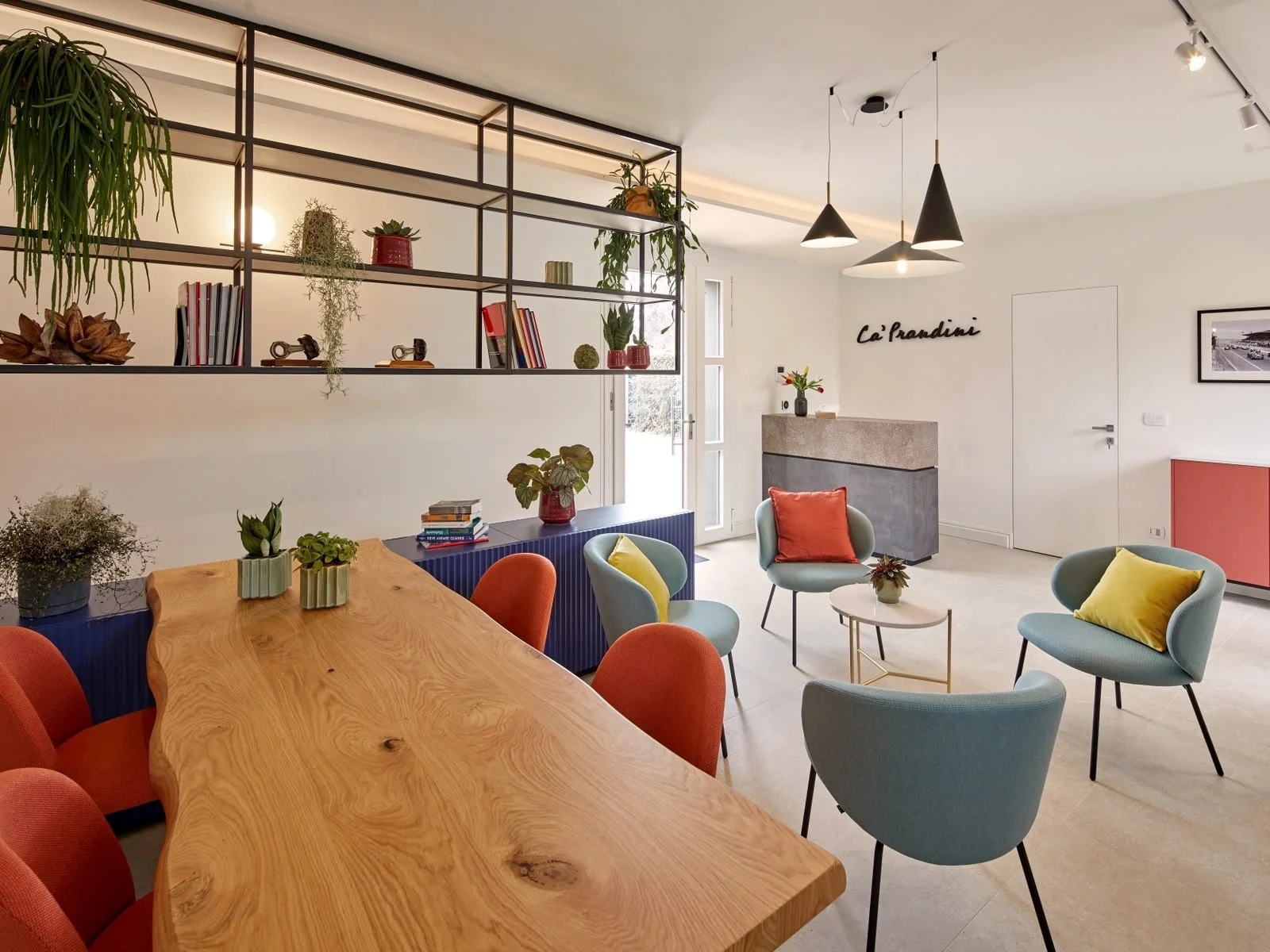 Modern interior of a living room or lounge area with a wooden table, colorful chairs and armchairs, decorative plants, books, and minimalist wall decor with the words 'Le Grandin' on the wall.