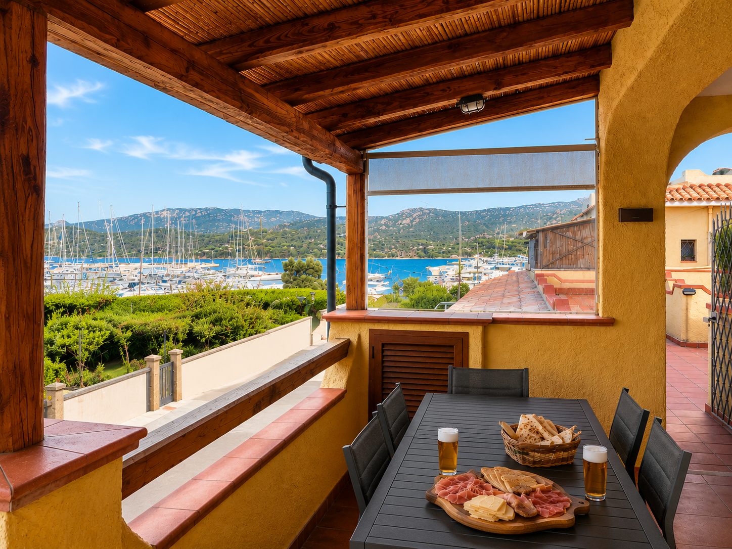 A cozy outdoor patio with a table set with food and drinks, overlooking a marina with boats and a scenic landscape of mountains and a blue sky.