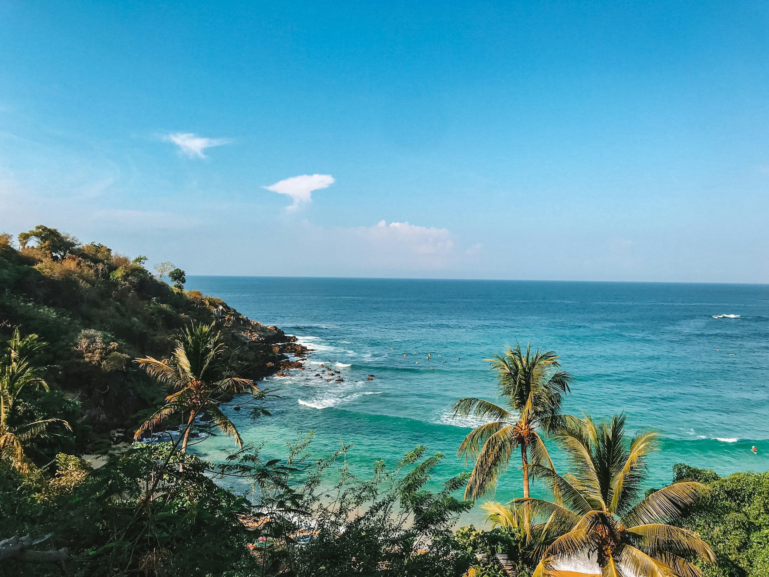 Tropical coastal scene with lush green trees and bushes on a cliff overlooking the ocean, clear turquoise water, and people swimming. Blue sky with a few clouds.
