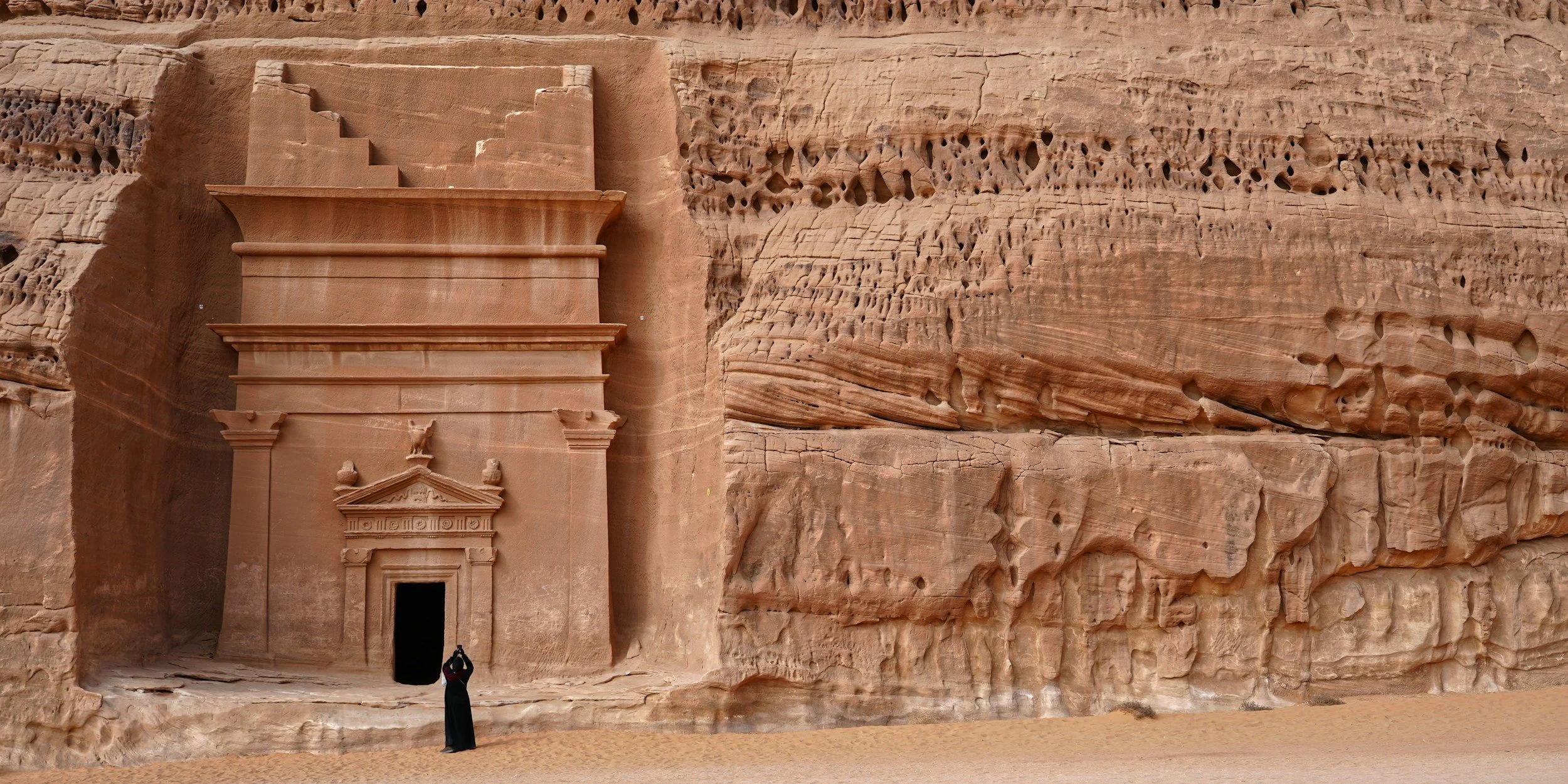 The image shows a rock-cut monument, Al-Khazneh, carved into a reddish sandstone cliff in Petra, Jordan. The facade features intricate architectural details, columns, and statues, set against a backdrop of rugged rock formations.