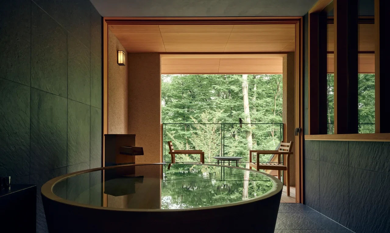 A modern bathroom with a dark bathtub and wooden accents, overlooking a green forest through a balcony.