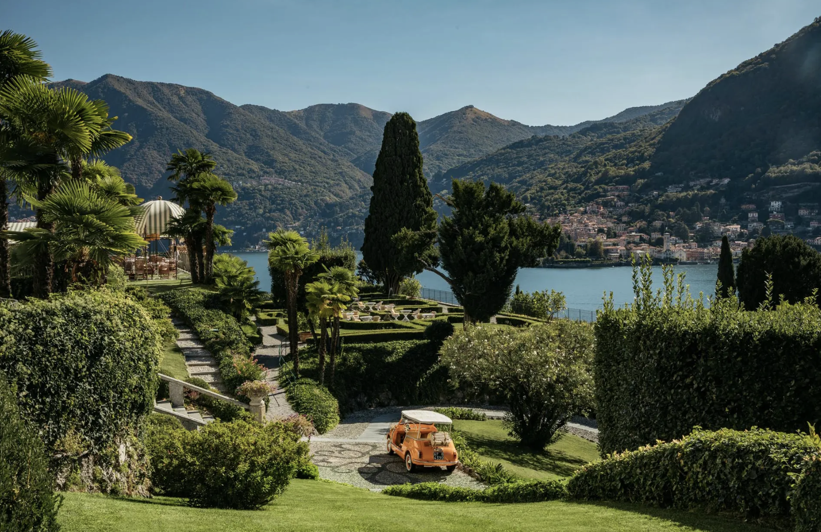 A lush, well-manicured garden with a vintage orange car parked on a stone driveway, overlooking a lake with mountains in the background. There are tall trees, hedges, and a gazebo with striped roof.