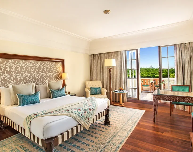 A hotel room with a large bed, armchair, floor lamp, window with curtains, and a balcony with outdoor furniture, overlooking greenery.