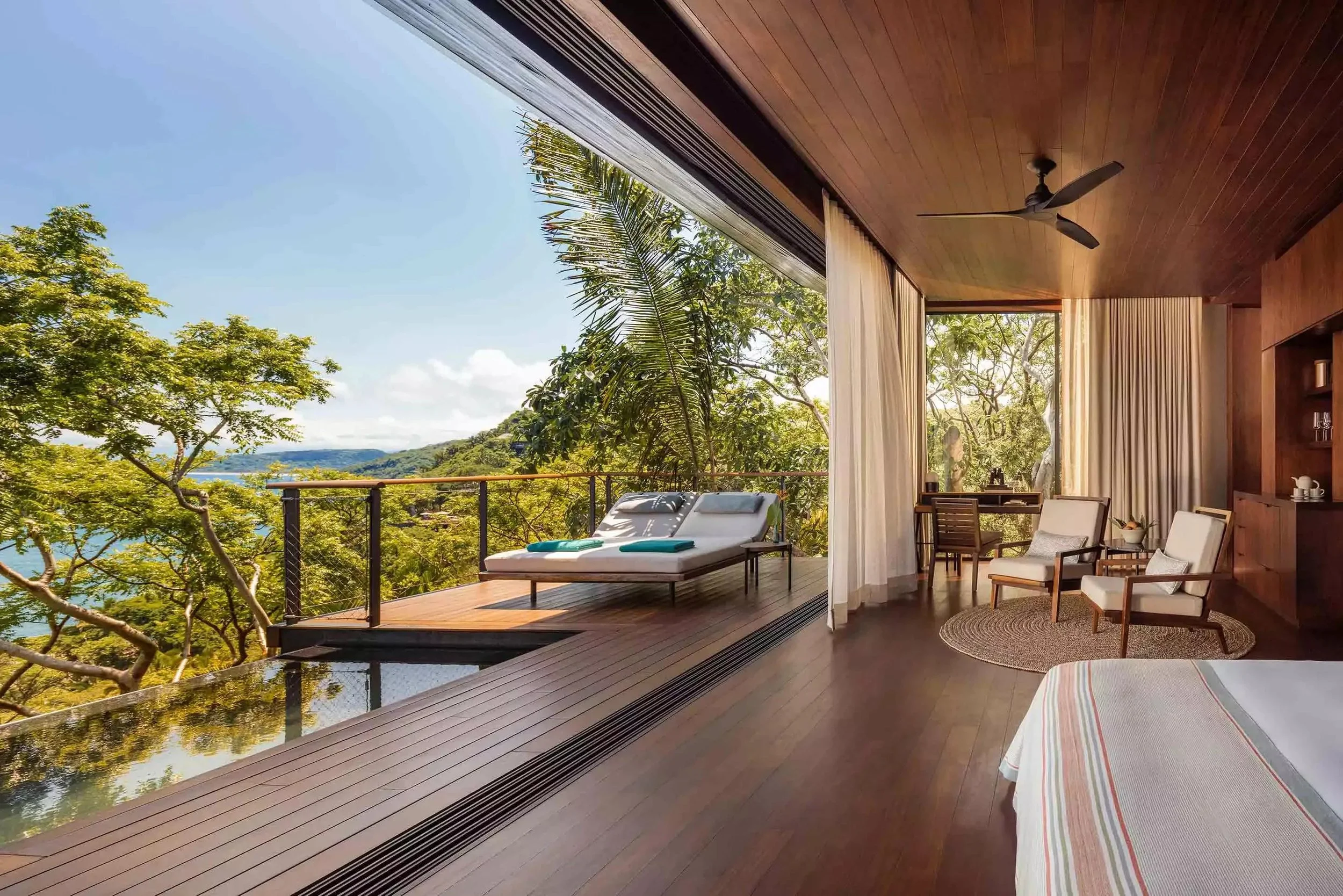 A cozy living space with large glass sliding doors open to a balcony surrounded by lush green trees and a scenic view of a body of water and hills in the distance. The room features wooden walls and ceiling, with chairs, a table, and a lounge chair on the balcony.