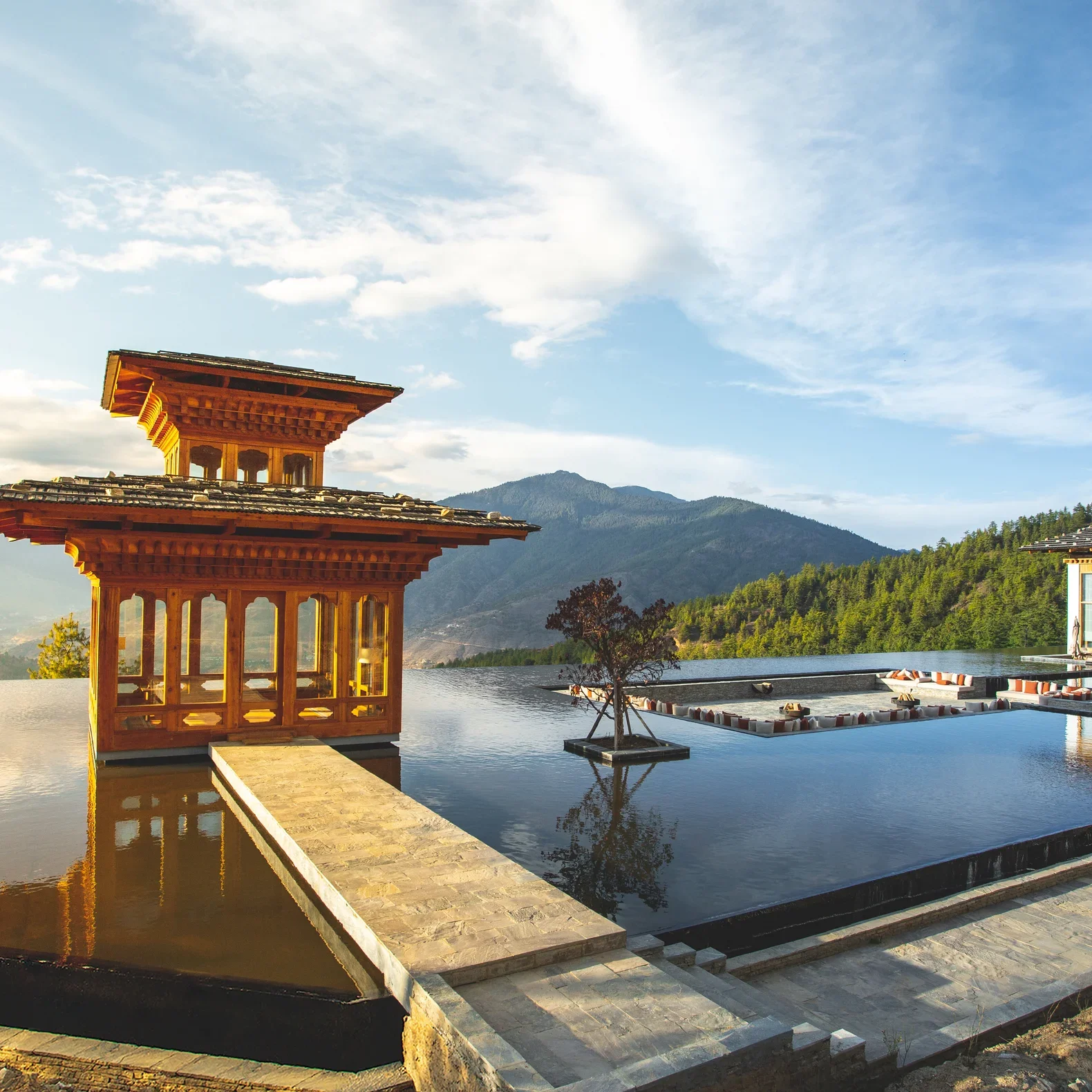 A scenic view of a wooden pagoda-style structure near a pool with a backdrop of mountains and a partly cloudy sky.