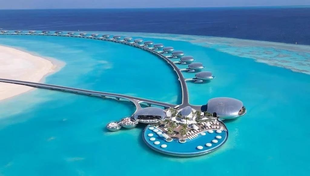 Aerial view of a luxury resort built on water with a circular pool area and overwater bungalows extending into the ocean.