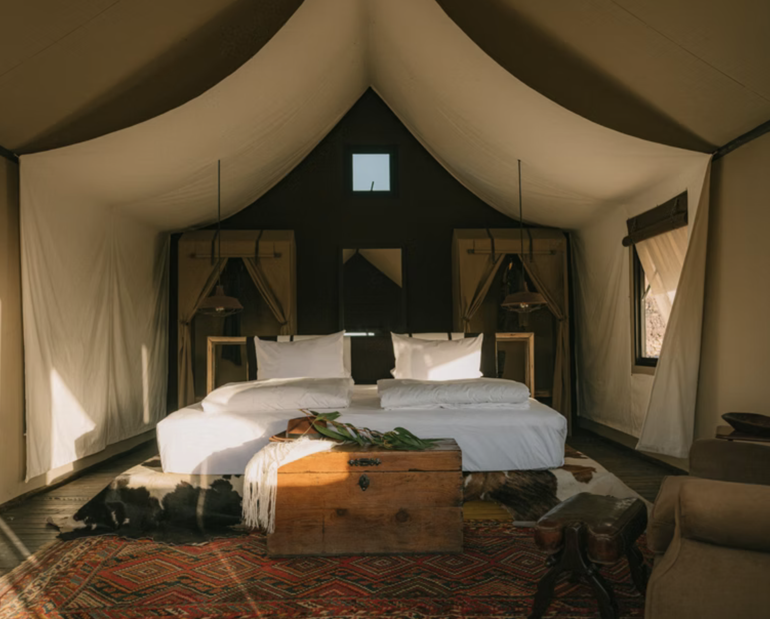 Interior of a cozy bedroom with a large bed, white linens, and wooden headboard, in a tent-like structure with draped fabric ceiling and windows on the sides, sunlight casting shadows.