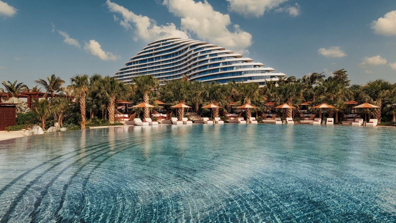A luxurious outdoor swimming pool with a modern, curved hotel building in the background, surrounded by palm trees and lounge chairs with umbrellas.