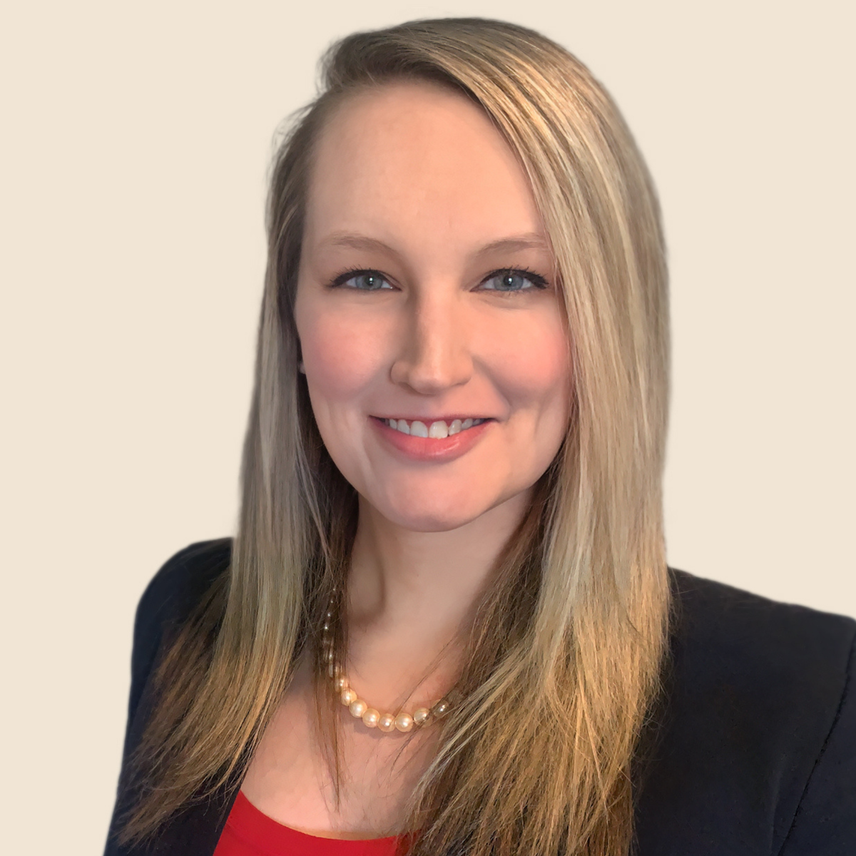 A professional woman with long blonde hair, blue eyes, wearing a black blazer, red top, and a pearl necklace, smiling against a plain light background.