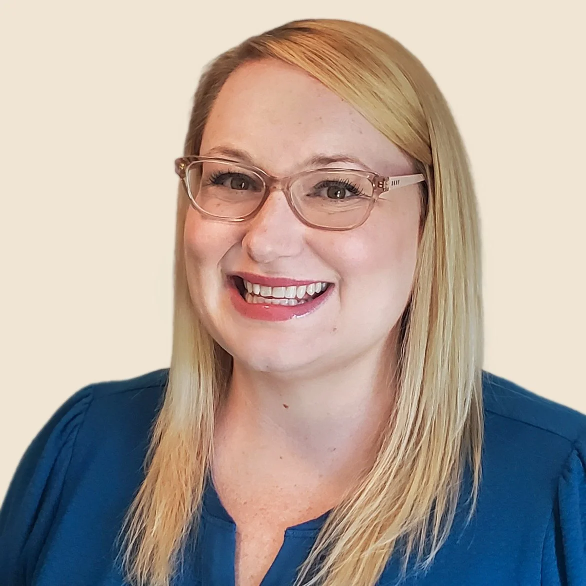 Close-up photo of a woman with blonde hair, glasses, smiling, wearing a blue top, against a plain background.