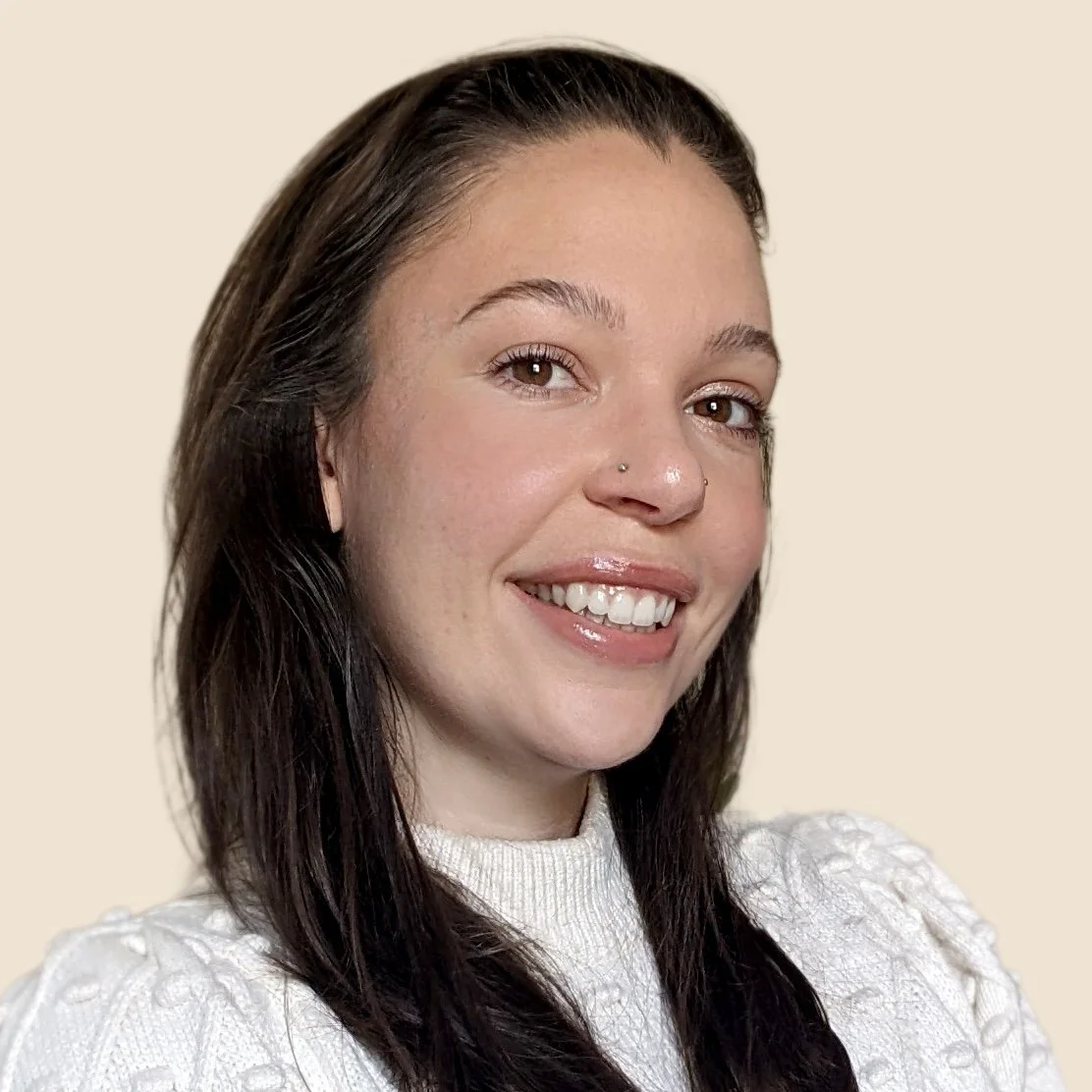 Close-up of a young woman with brown hair, brown eyes, and light skin, smiling and wearing a white knitted sweater against a beige background.