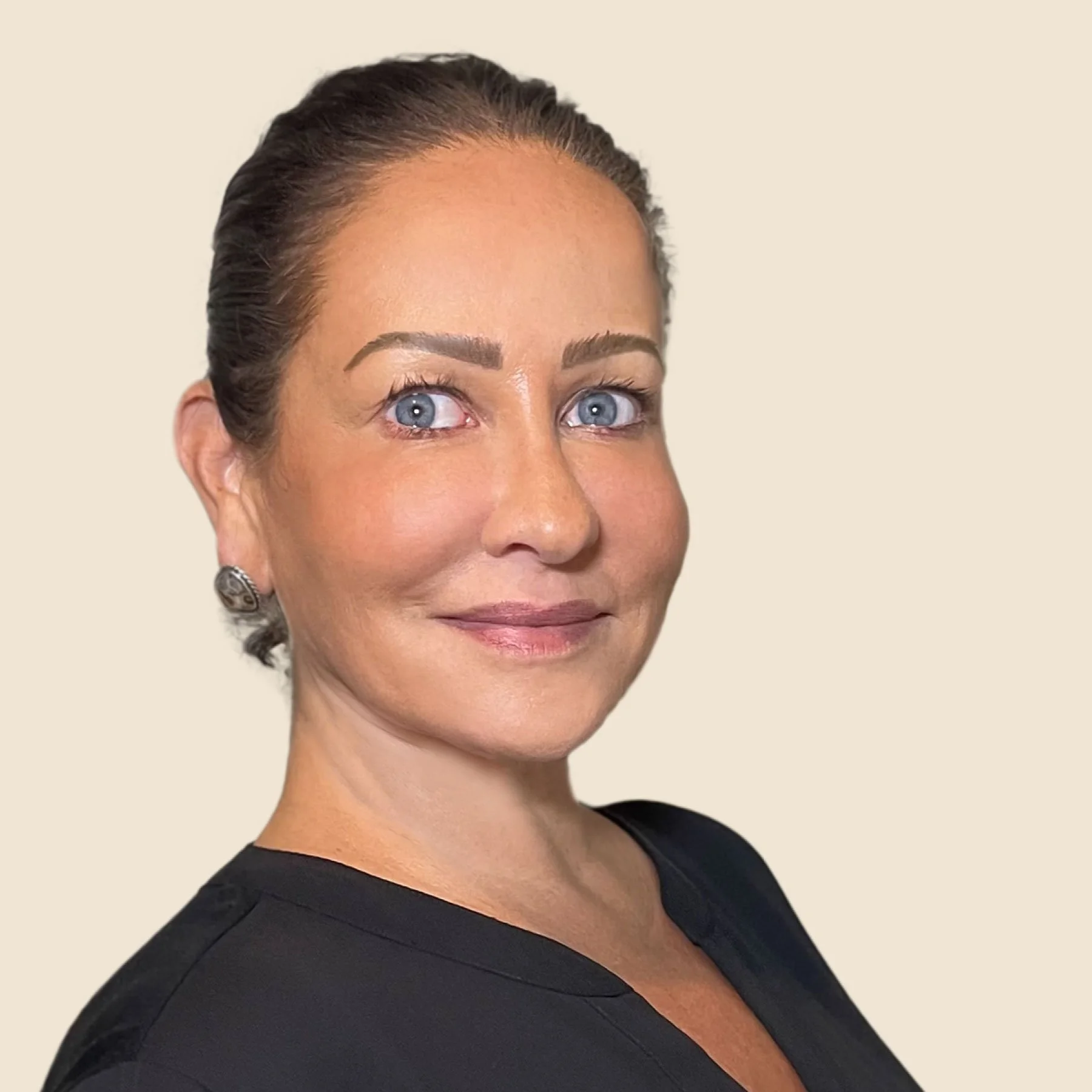 Close-up of a woman with light skin, blue eyes, dark hair pulled back, wearing a black top, and silver earrings, smiling softly against a plain beige background.