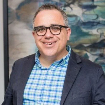 A man wearing glasses, a blue checkered shirt, and a dark blazer, smiling in front of an abstract background.