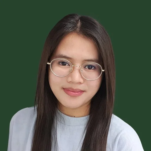 Portrait of a young woman with long dark hair and glasses, wearing a light-colored top, standing against a green background.