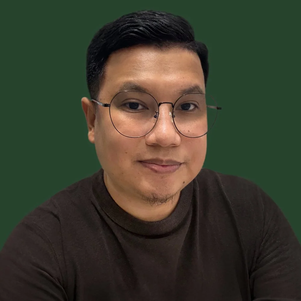 A young man with glasses and short black hair, wearing a black shirt, looking at the camera with a slight smile, against a solid dark green background.