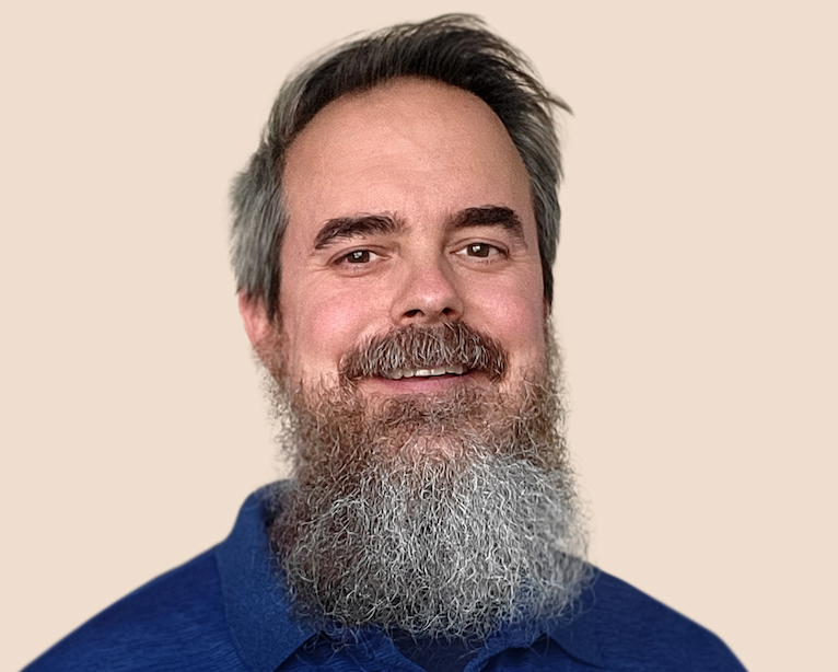 Portrait of a smiling middle-aged man with a gray beard and hair, wearing a blue shirt, against a plain beige background.