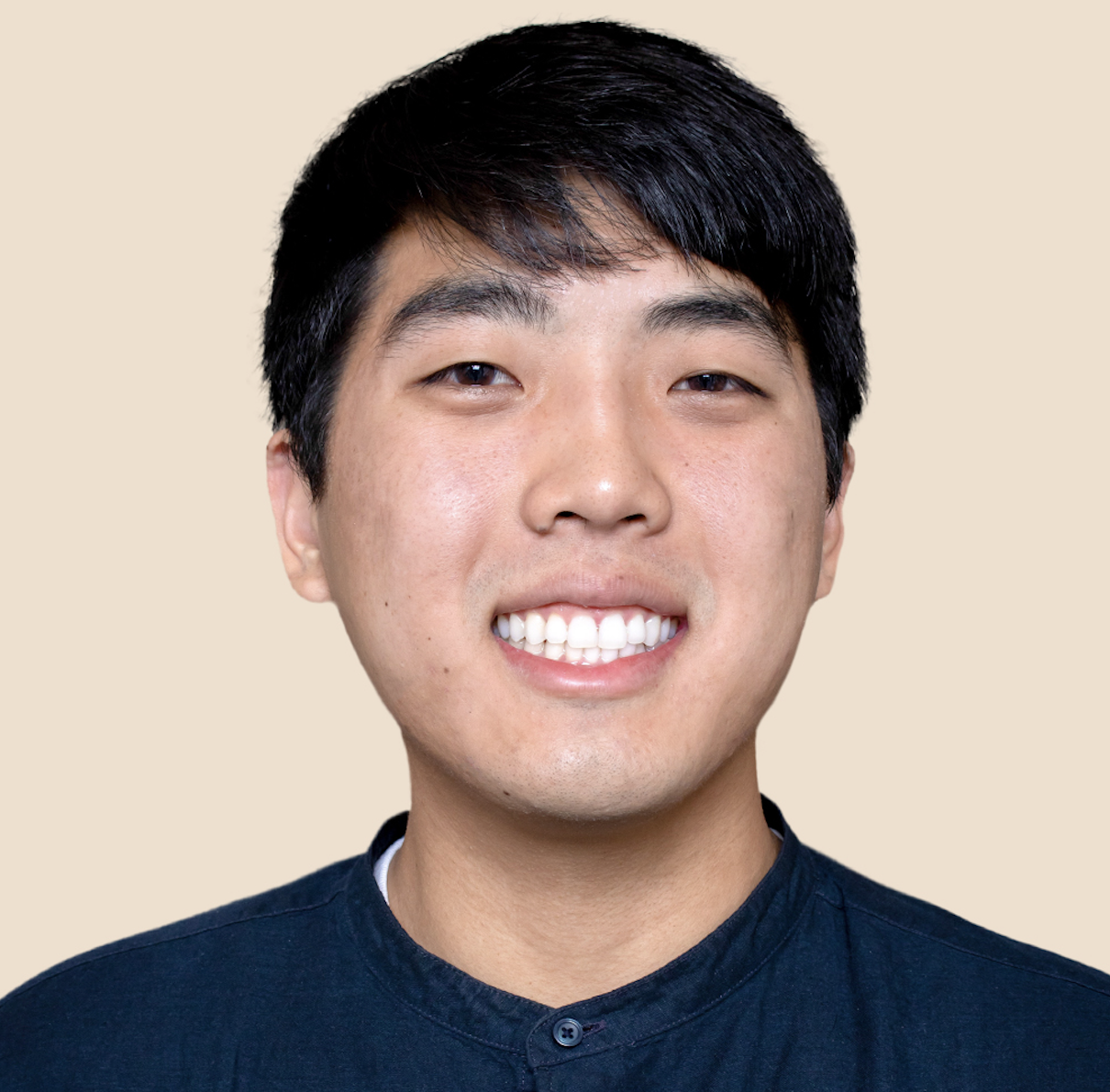 A young Asian man with black hair and a wide smile showing his teeth, wearing a dark shirt, against a beige background.
