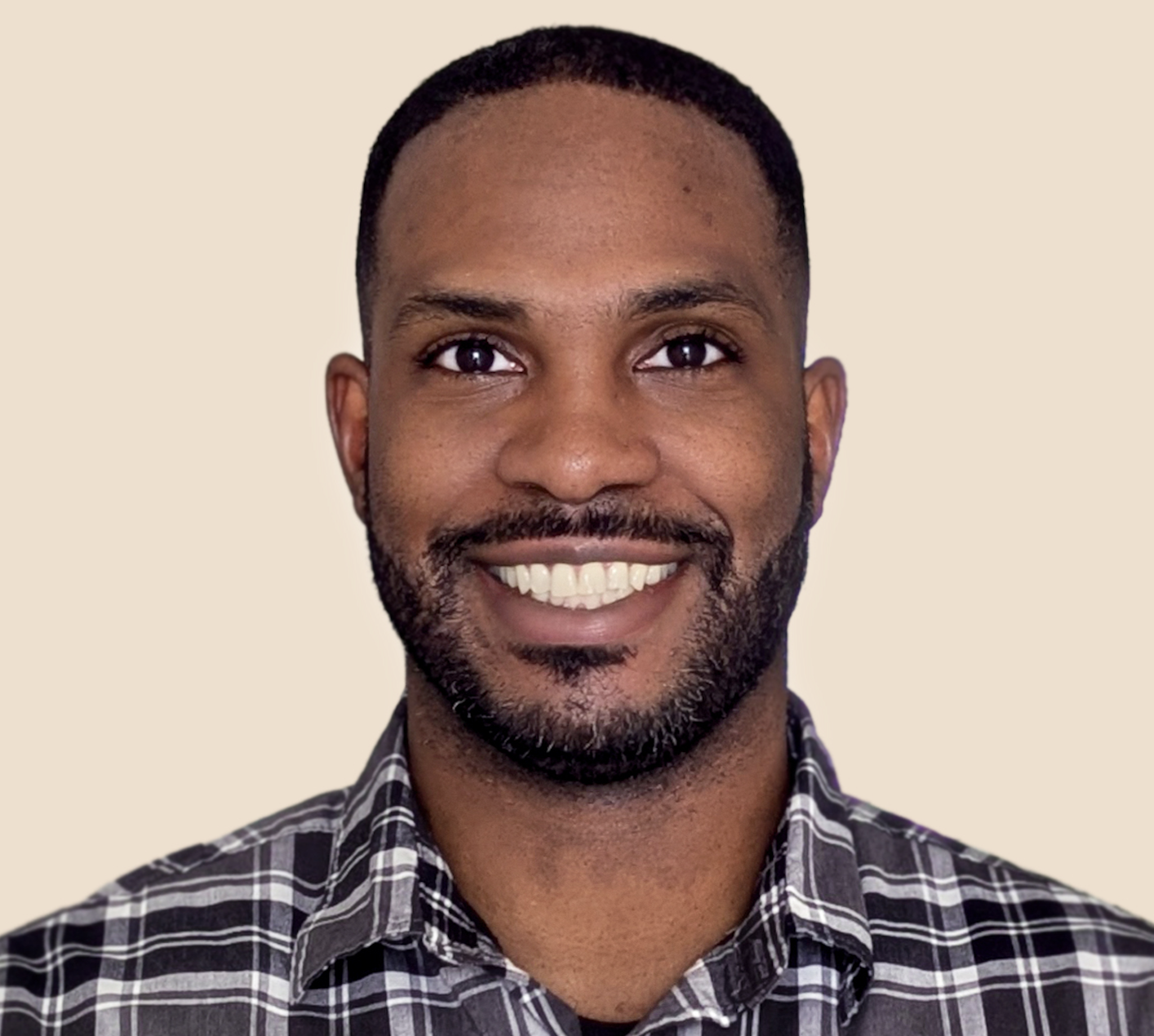 Portrait of a smiling man with a short beard and mustache, wearing a black and white checkered shirt, against a plain light beige background.