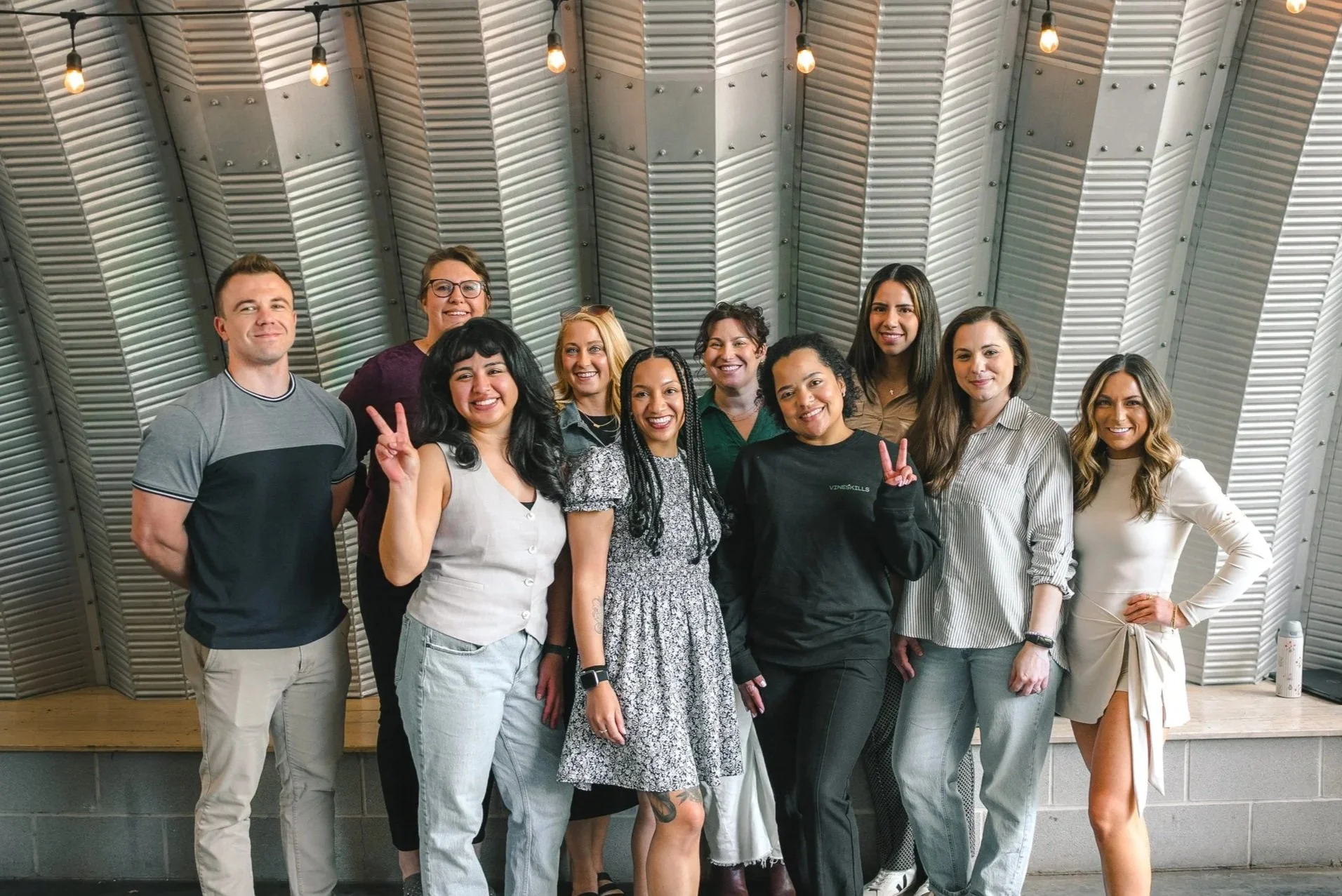 Group of ten diverse people, women and men, standing together in a modern industrial-style room with metallic wall panels and exposed light bulbs, smiling and posing for a photo.