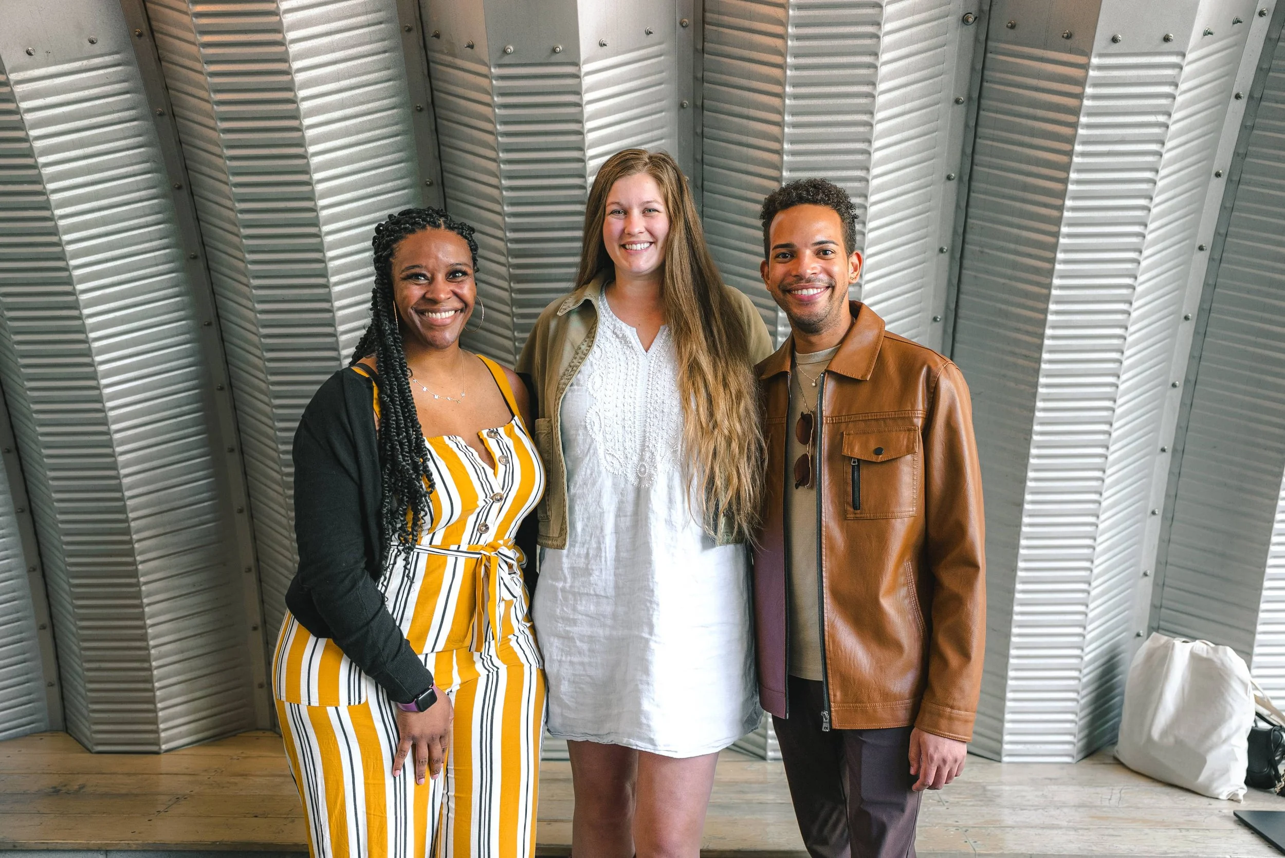 Three people standing together in front of a metallic, ribbed wall, smiling at the camera. The person on the left is an African American woman with braids, wearing a yellow, white, and black striped dress with a black cardigan. The person in the middle is a woman with long, wavy brown hair, wearing a white dress and a tan jacket. The person on the right is a man with short curly hair, wearing a brown leather jacket and a beige shirt.
