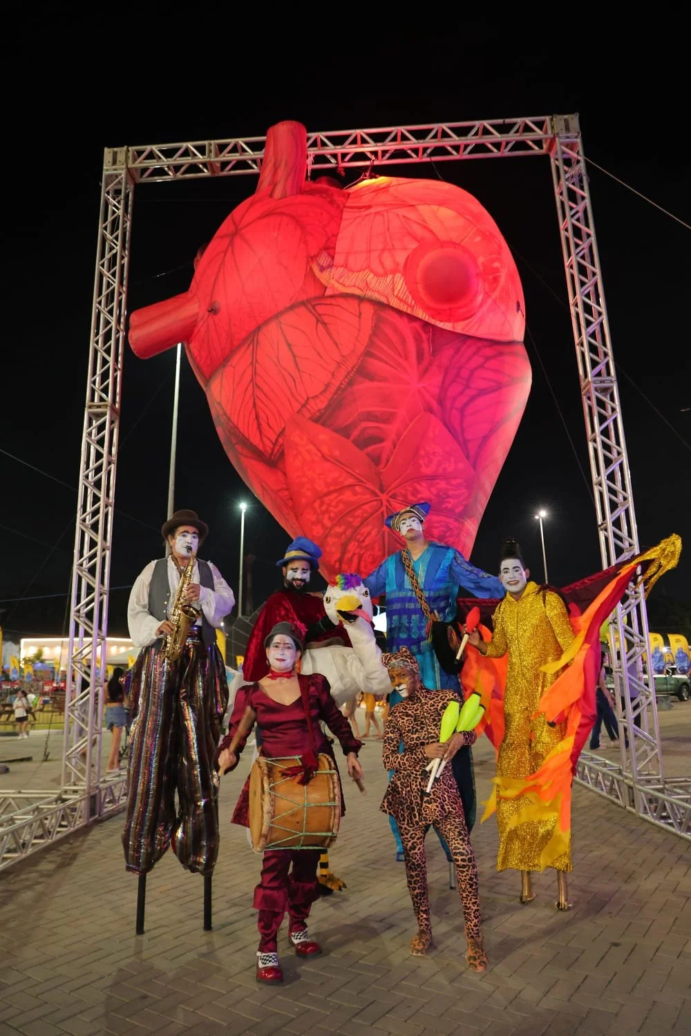 Grupo de artistas de circo com maquiagem de palhaço, usando fantasias coloridas, posando na frente de uma escultura gigante de coração iluminada, durante a noite em um parque de diversões.