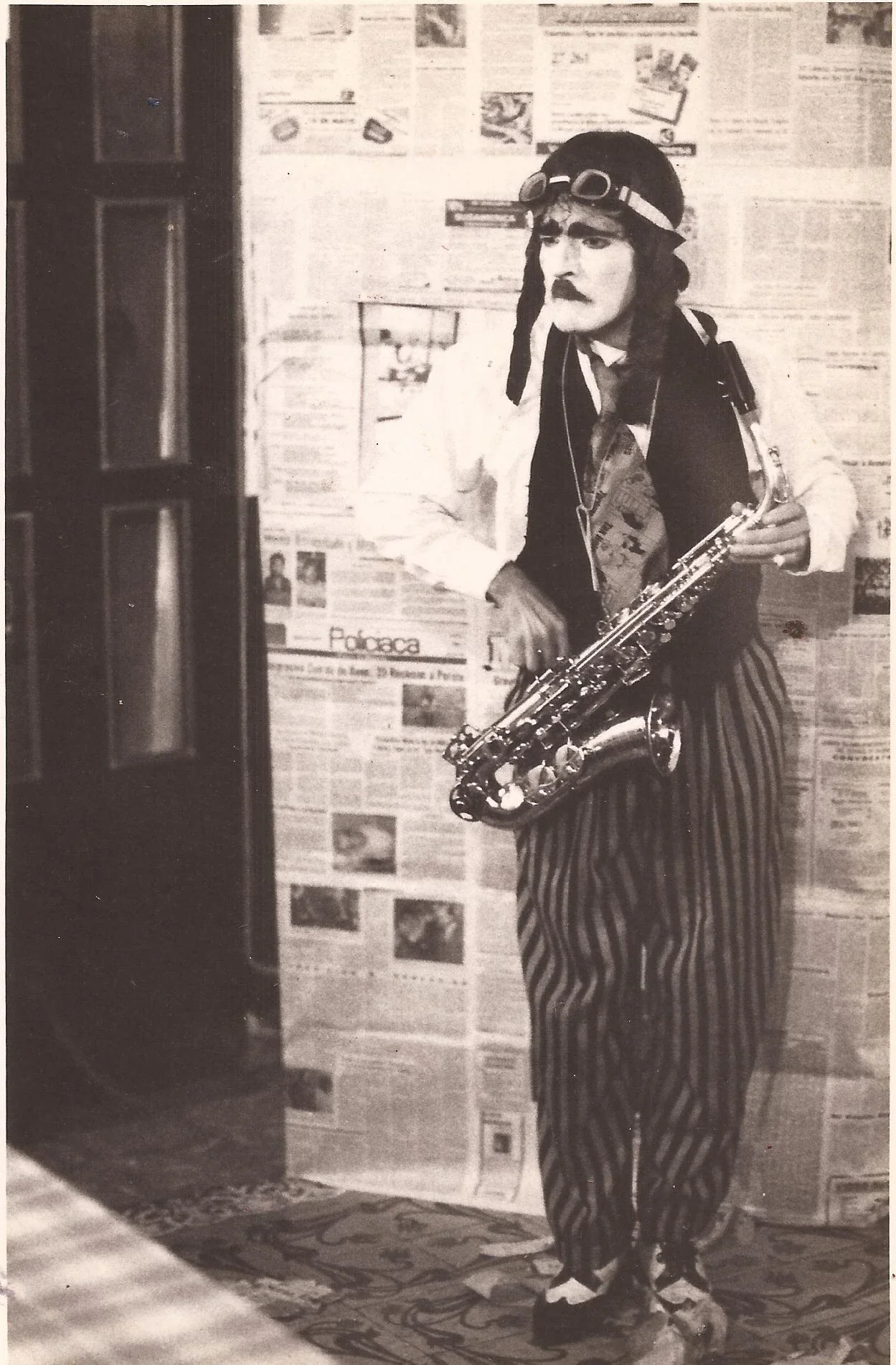 Homem vestido de palhaço ou artista com roupas listradas, chapéu, óculos e bigode, segurando um saxofone, em ambiente decorado com jornais na parede.