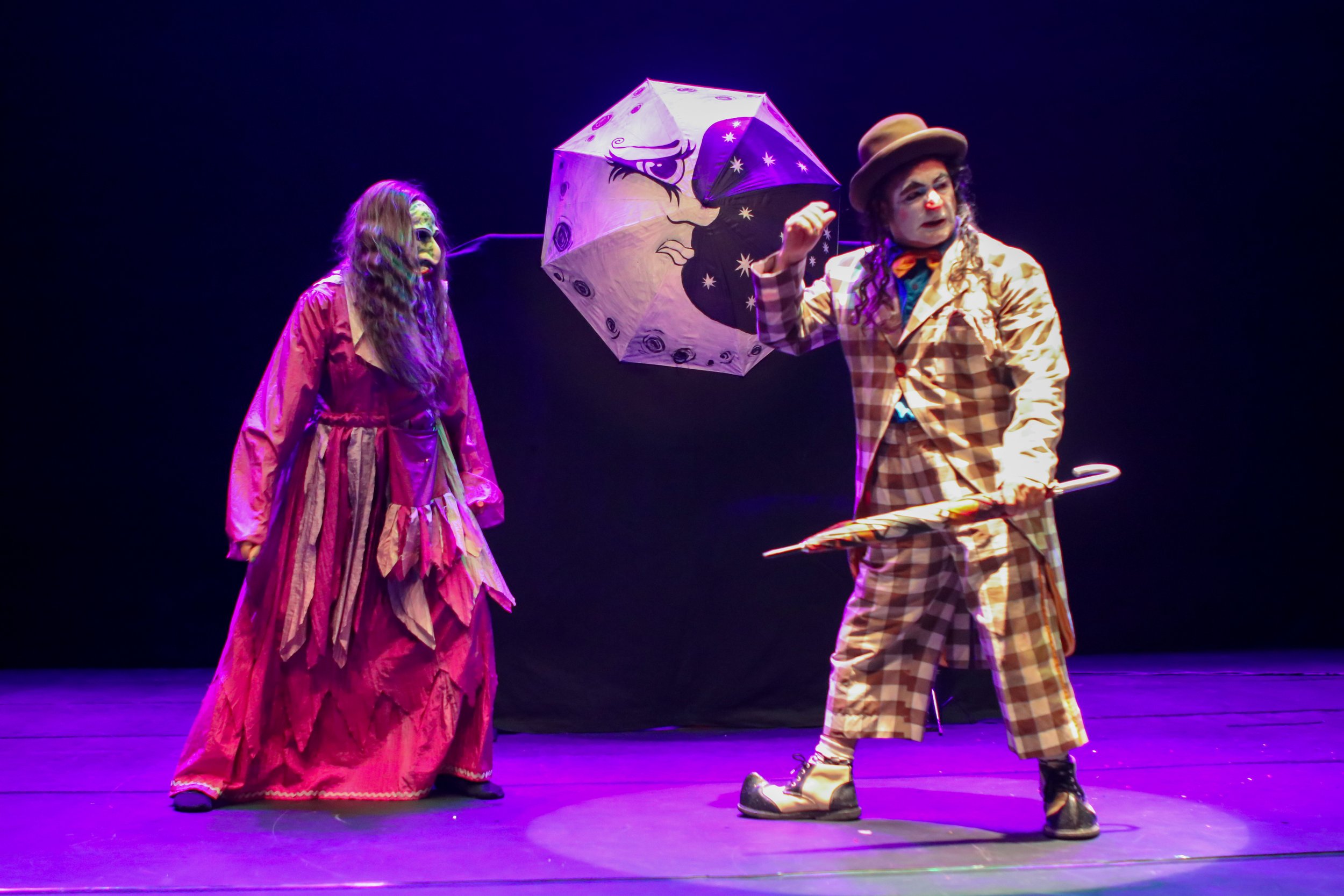 Dois atores de teatro performando, um com maquiagem e vestido colorido, outro com roupas de abuclha, guarda-chuva decorado com estrelas e lua, em palco com iluminação rosa e roxa.