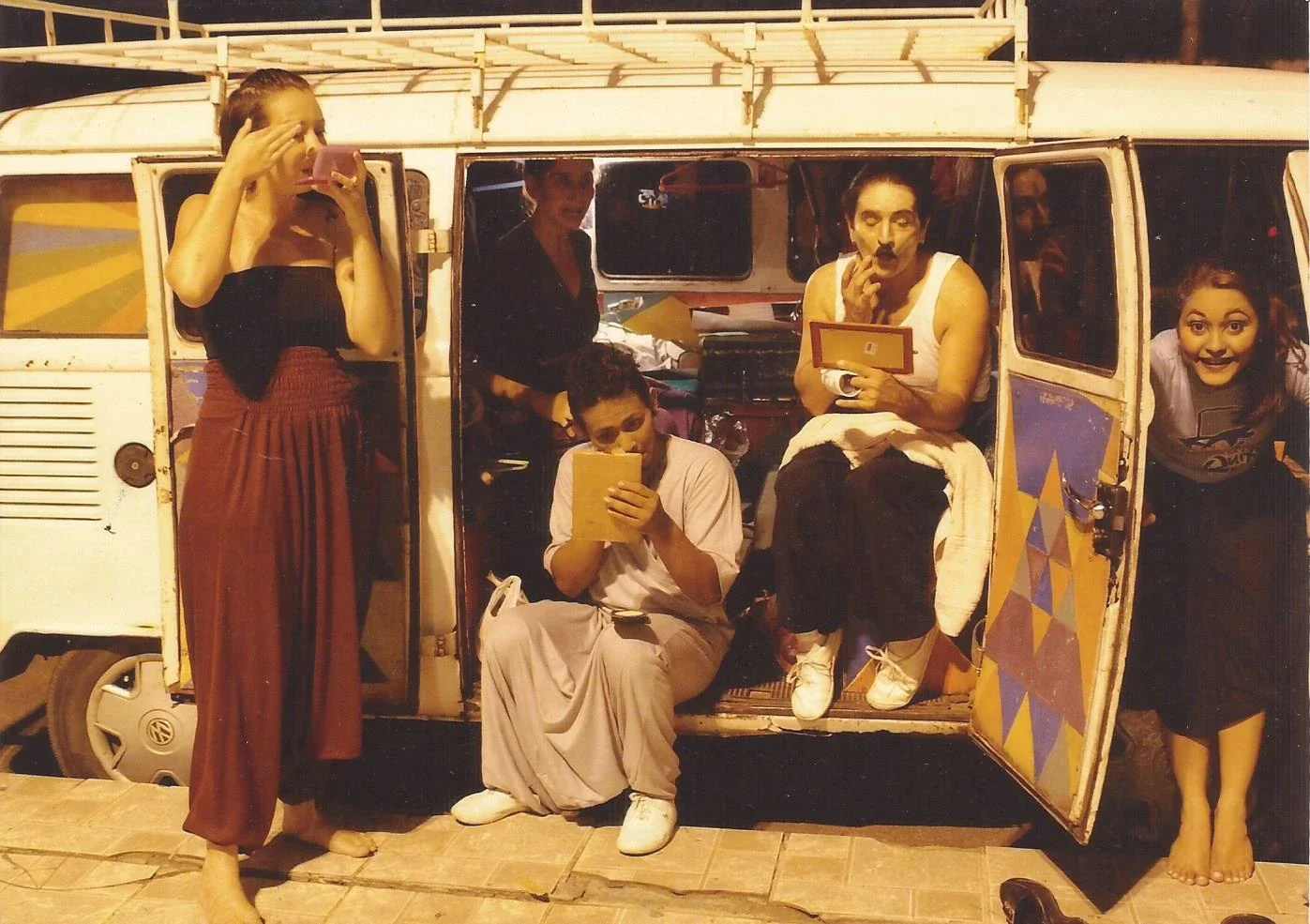 Grupo de pessoas com figurinos característicos, posando ao redor de uma van decorada, em uma situação que parece ser uma apresentação ou evento, durante a noite.
