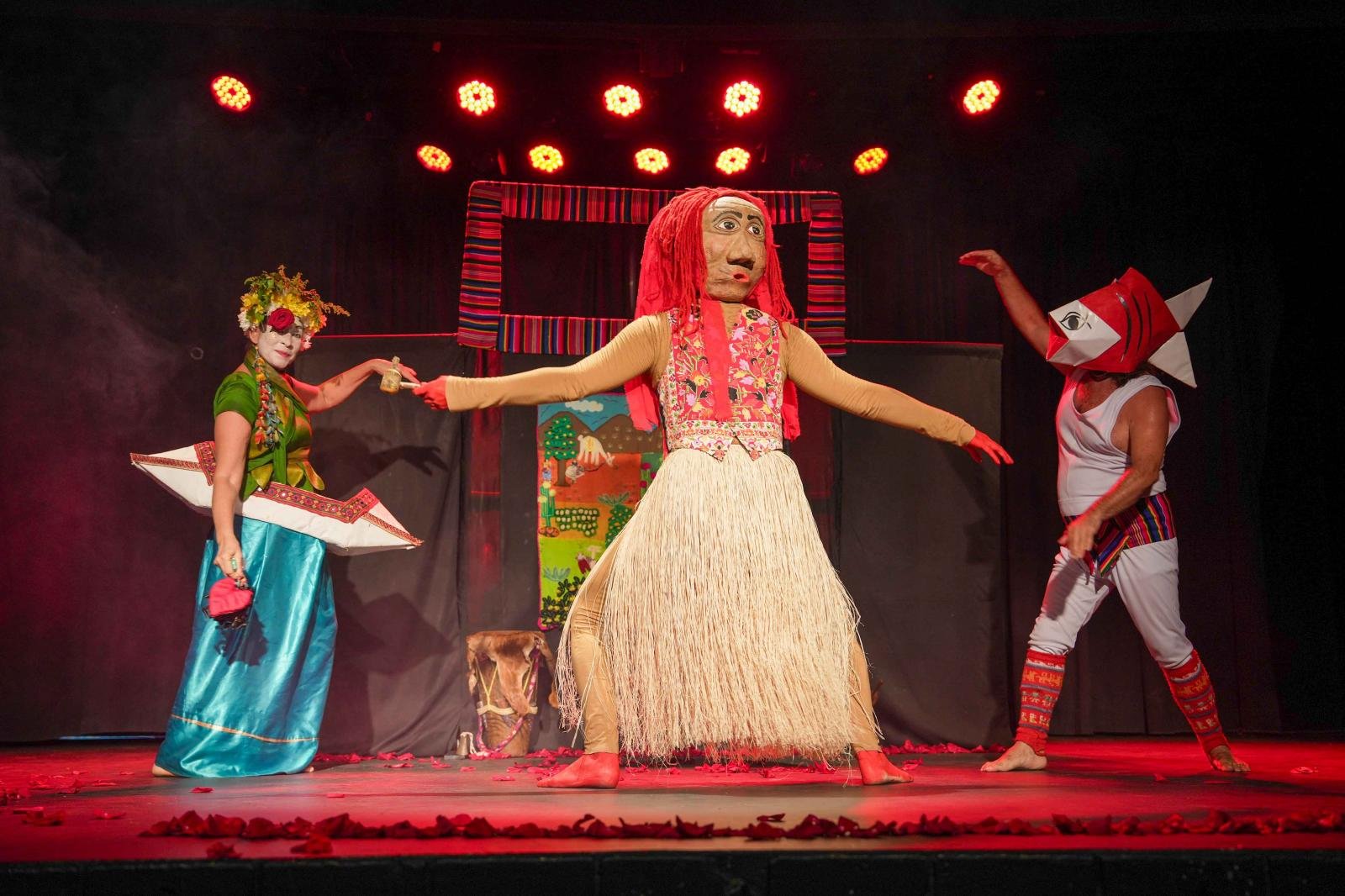 Cena de uma peça de teatro com três atores caracterizados com roupas coloridas e máscaras elaboradas. Um ator com máscara de rosto grande, vestindo saia de palha, está no centro, enquanto os outros dois, com fantasias ornamentadas, estão de cada lado e parecem interagir com ele, em um palco iluminado com luzes vermelhas.