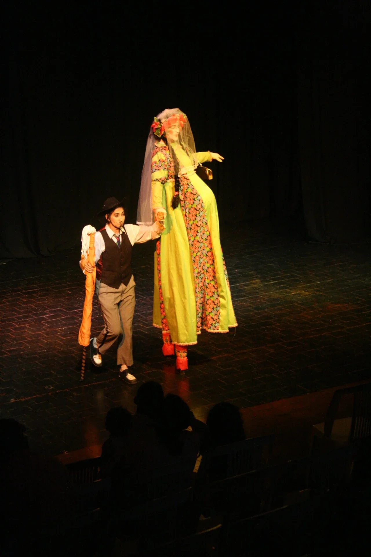 Duas pessoas em cena teatral, uma em um figurino colorido e alta, a outra menor e vestida com roupas formais, em palco com fundo escuro.