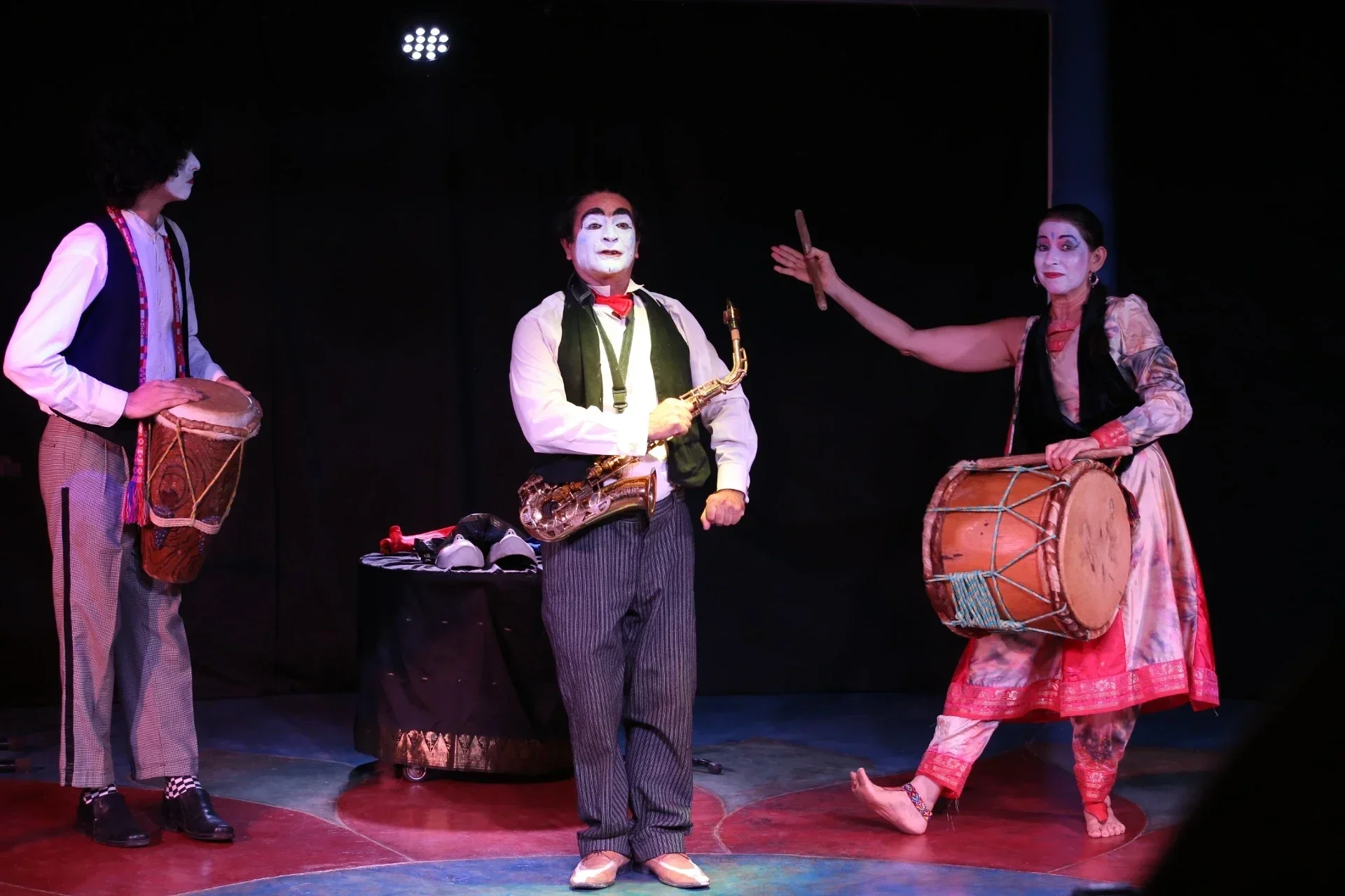 Cena de uma apresentação teatral com três atores com maquiagem de palhaço, usando roupas coloridas, em um palco escuro. Um deles segura um saxofone, outro tem um tambor, e o terceiro está com um pandeiro.