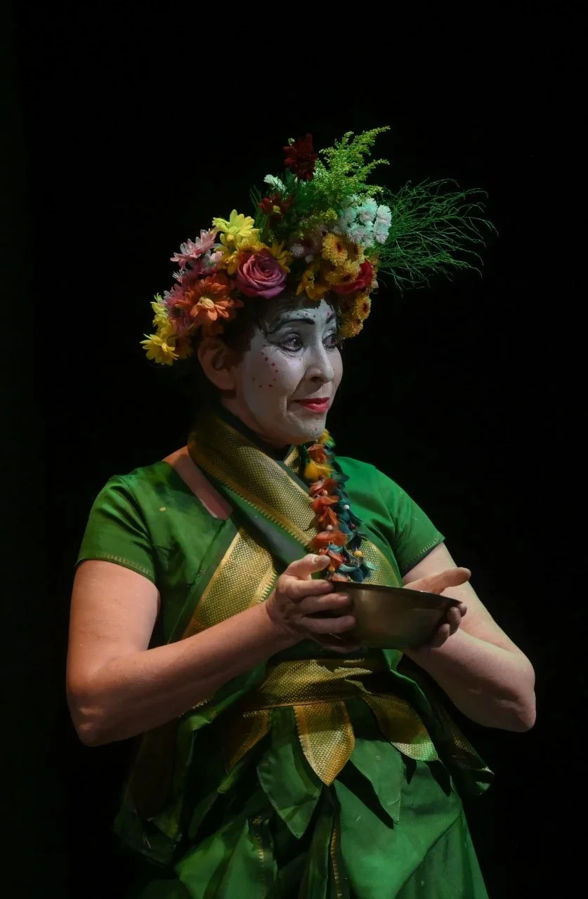 Mulher idosa com maquiagem de palhaço, usando vestido verde e acessório de flor, segurando uma tigela, contra fundo preto.