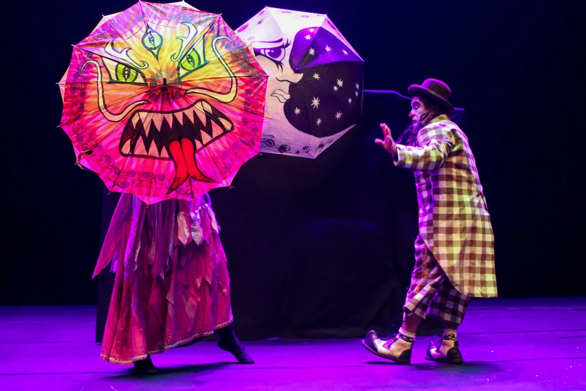 Duas pessoas de palhaço realizando uma performance com guarda-chuvas decorados; uma veste uma roupa rosa de chifres e a outra uma fantasia de xadrez com chapéu marrom.