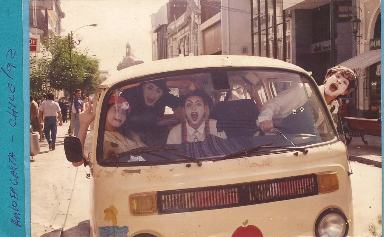 Grupo de pessoas em um micro-ônibus vintage, com expressões animadas, na rua de uma cidade antiga, com edifícios históricos ao fundo, causando nostalgia.