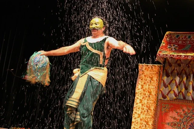 Boneca ou personagem de teatro com barba, vestida com traje tradicional, sob chuva teatral, com pano decorativo ao fundo
