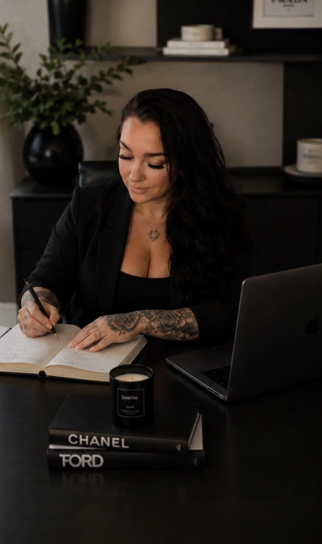 A woman with dark hair and tattoos on her arms writing in a notebook at a desk, with a MacBook, a candle, and two books titled 'Chanel' and 'Ford' in front of her.