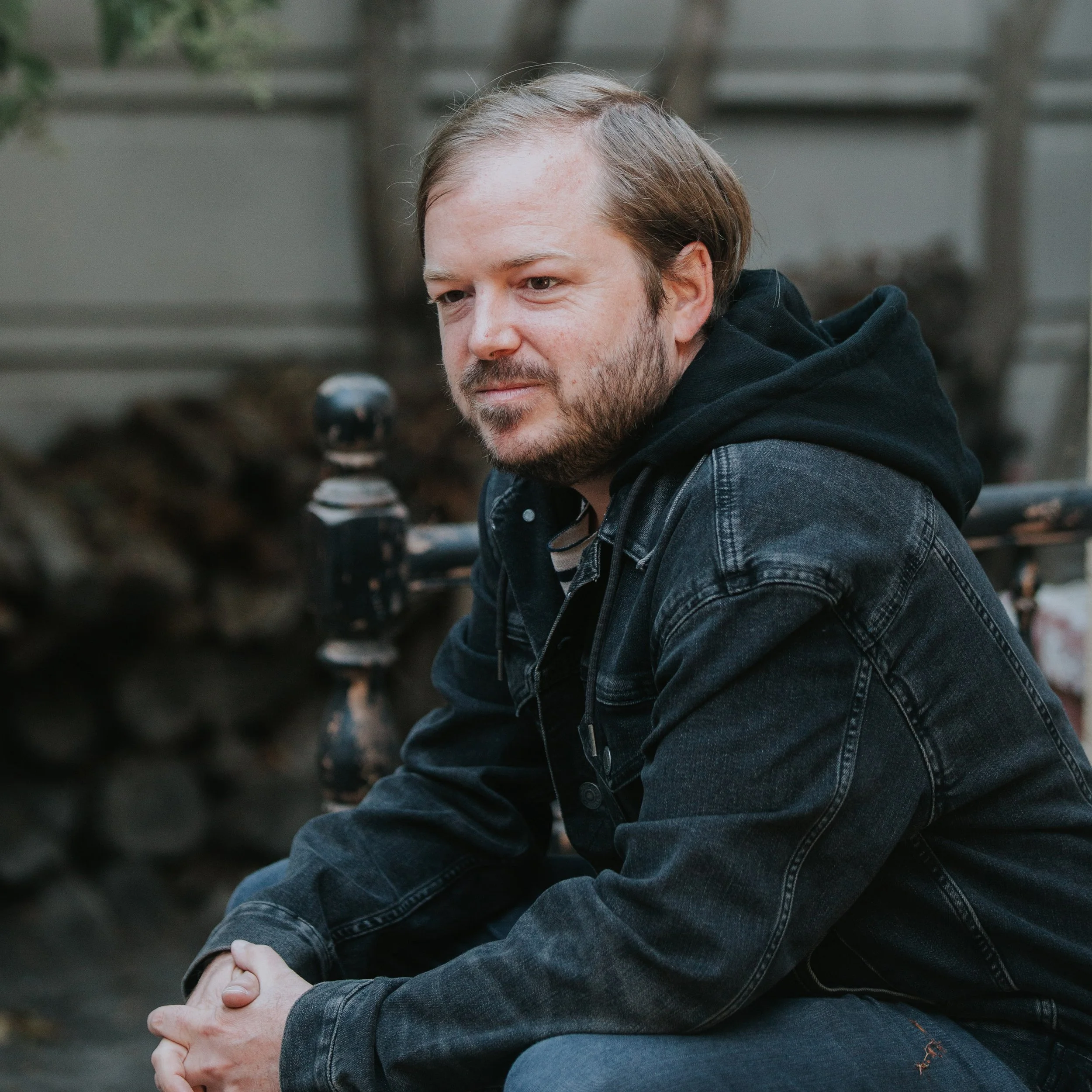 A close-up of the author, John T. Trautman, with light skin, brown hair, and a beard, sitting outdoors with trees and a wooden fence in the background. He is wearing a black hoodie and a denim jacket, looking slightly down and to the side.