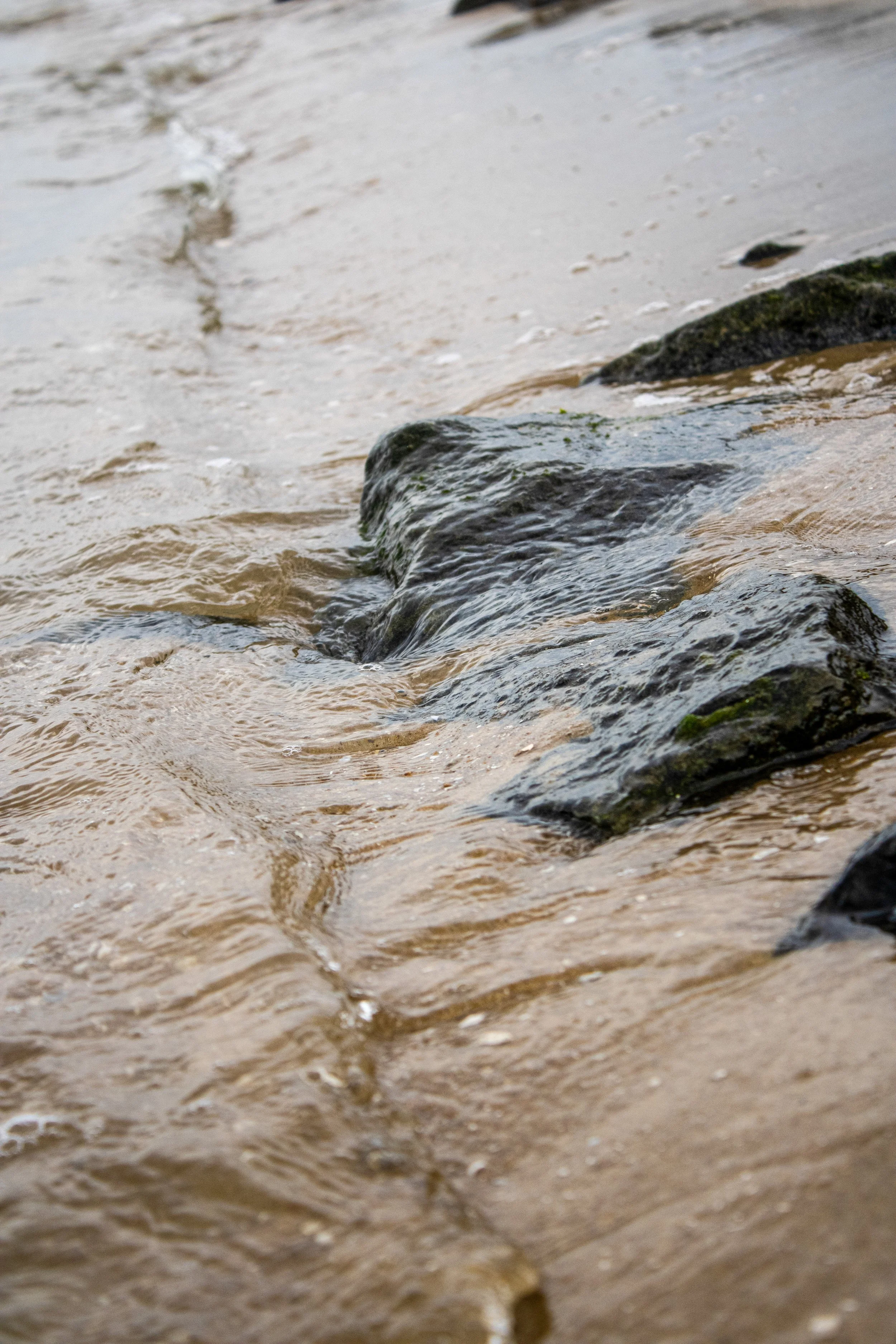 Close-up of sand and rocks at the edge of a body of water, with gentle waves washing over the rocks.
