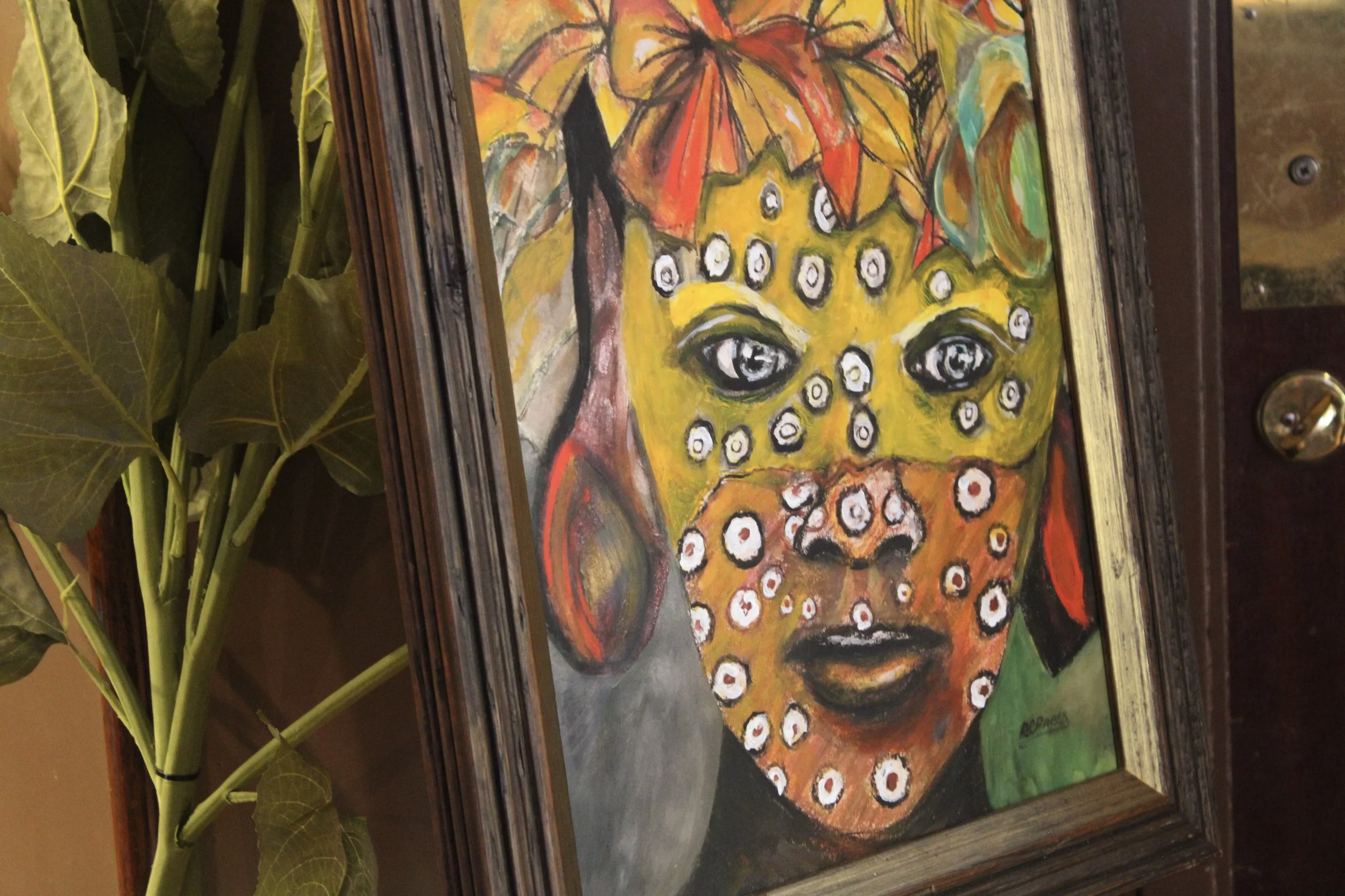 Colorful abstract painting of a face with prominent eyes, surrounded by orange and red shapes, framed in dark wood, with green foliage to the side.