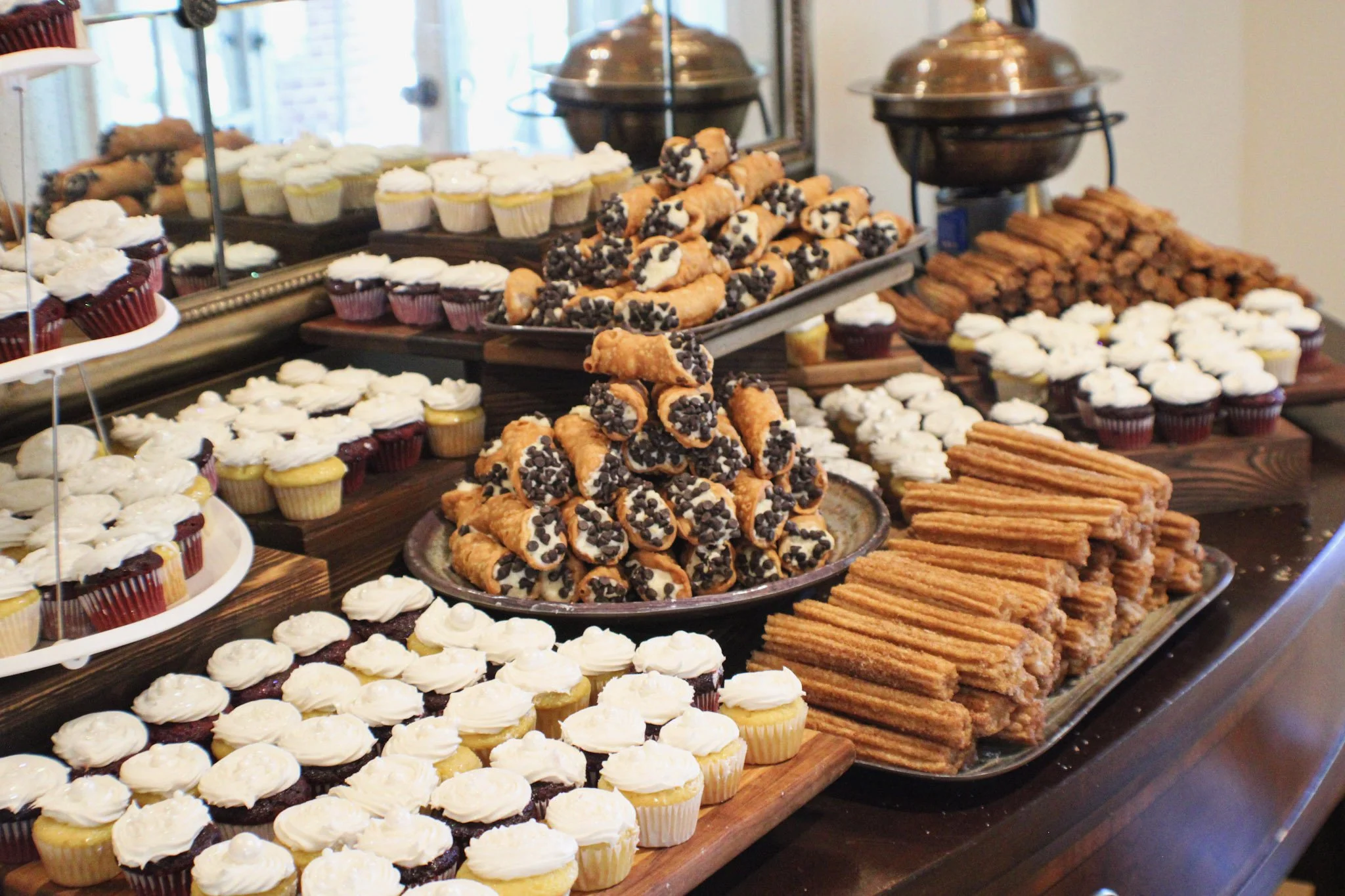 A display of assorted desserts including cupcakes, cannoli, churros with chocolate chips, cookies, and layered pastries on a large wooden table.