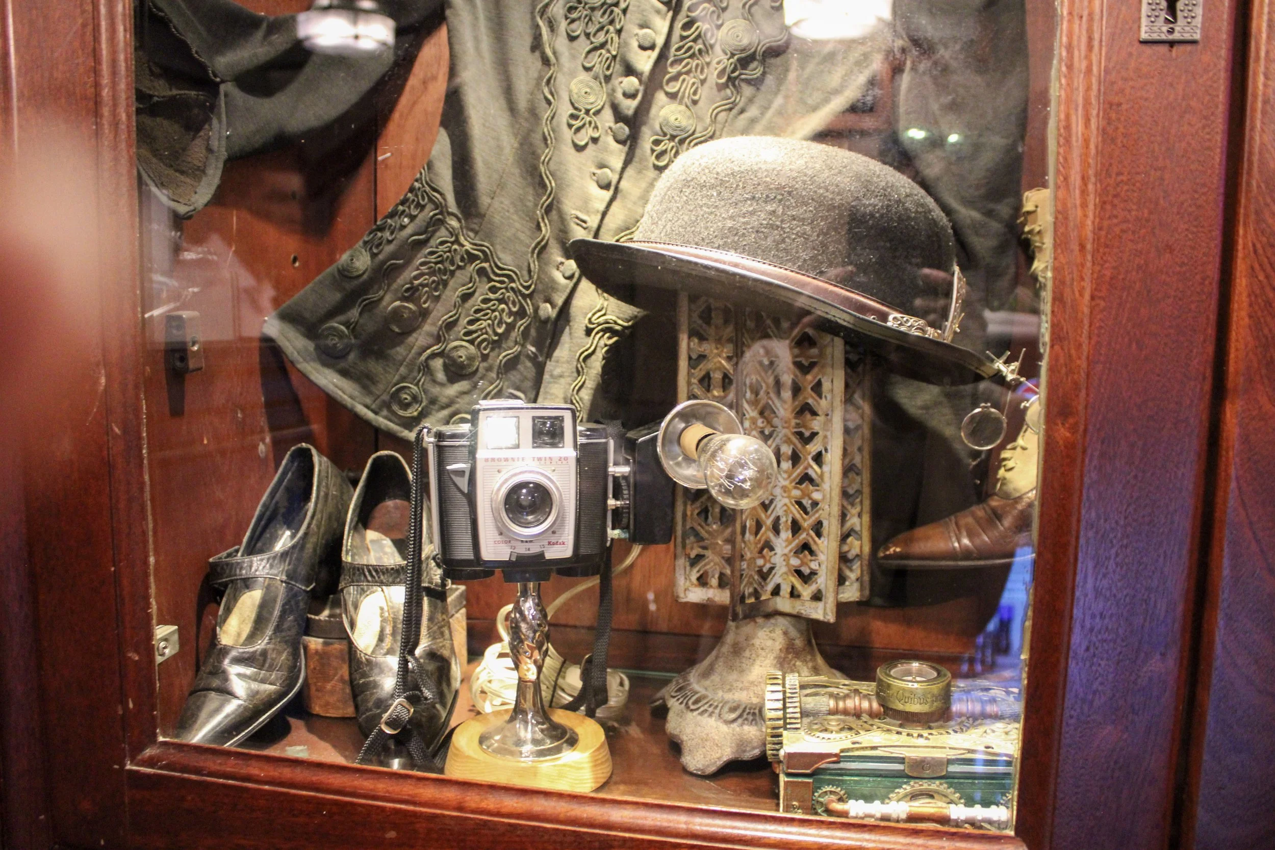 Display case containing vintage items including black vintage shoes, a grey fedora hat, an old camera, a brass key, a candle, and other miscellaneous objects.