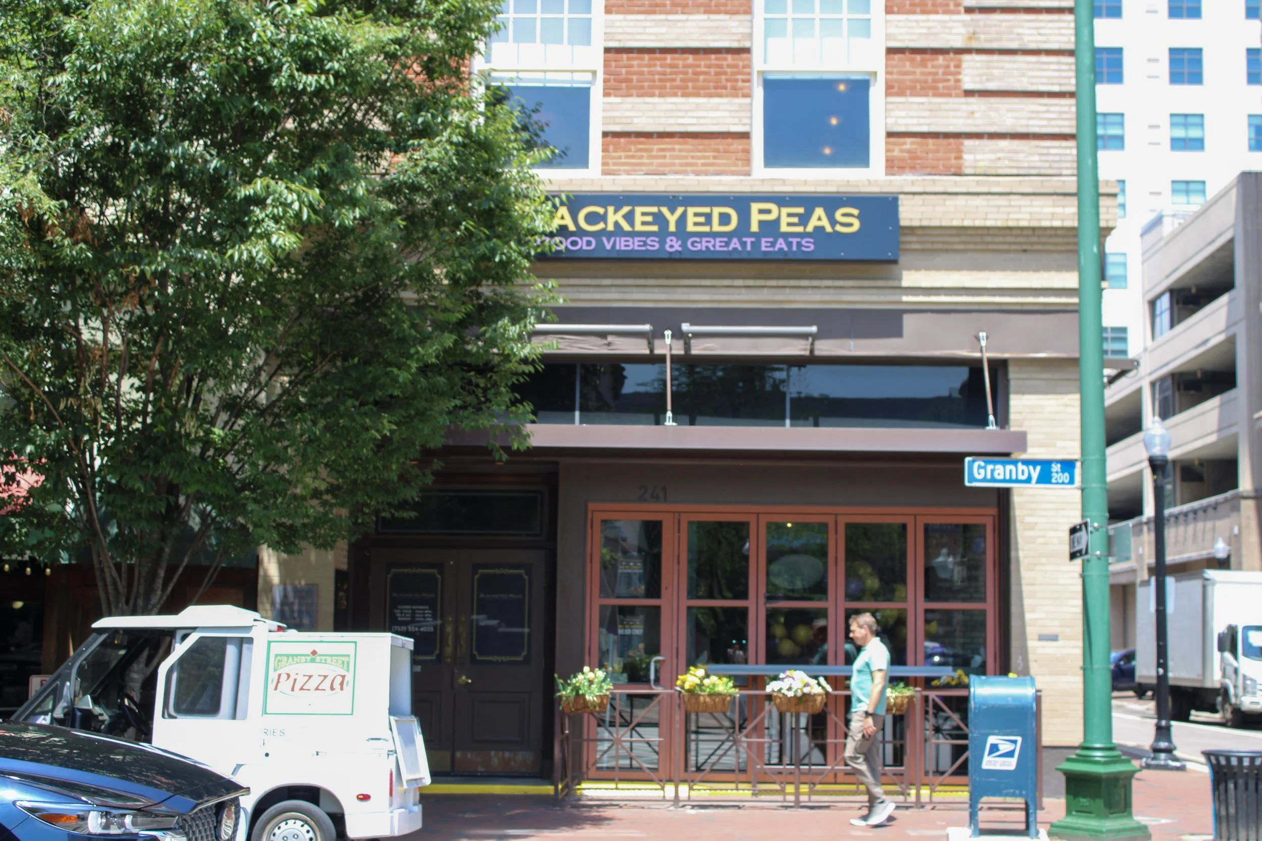 Street view with a restaurant named 'Backeyed Peas' with a sign that says 'Food Vibes & Great Eats.' A man is walking past the storefront which has large windows and red doors. There are trees, cars, street signs, and a blue mailbox nearby.