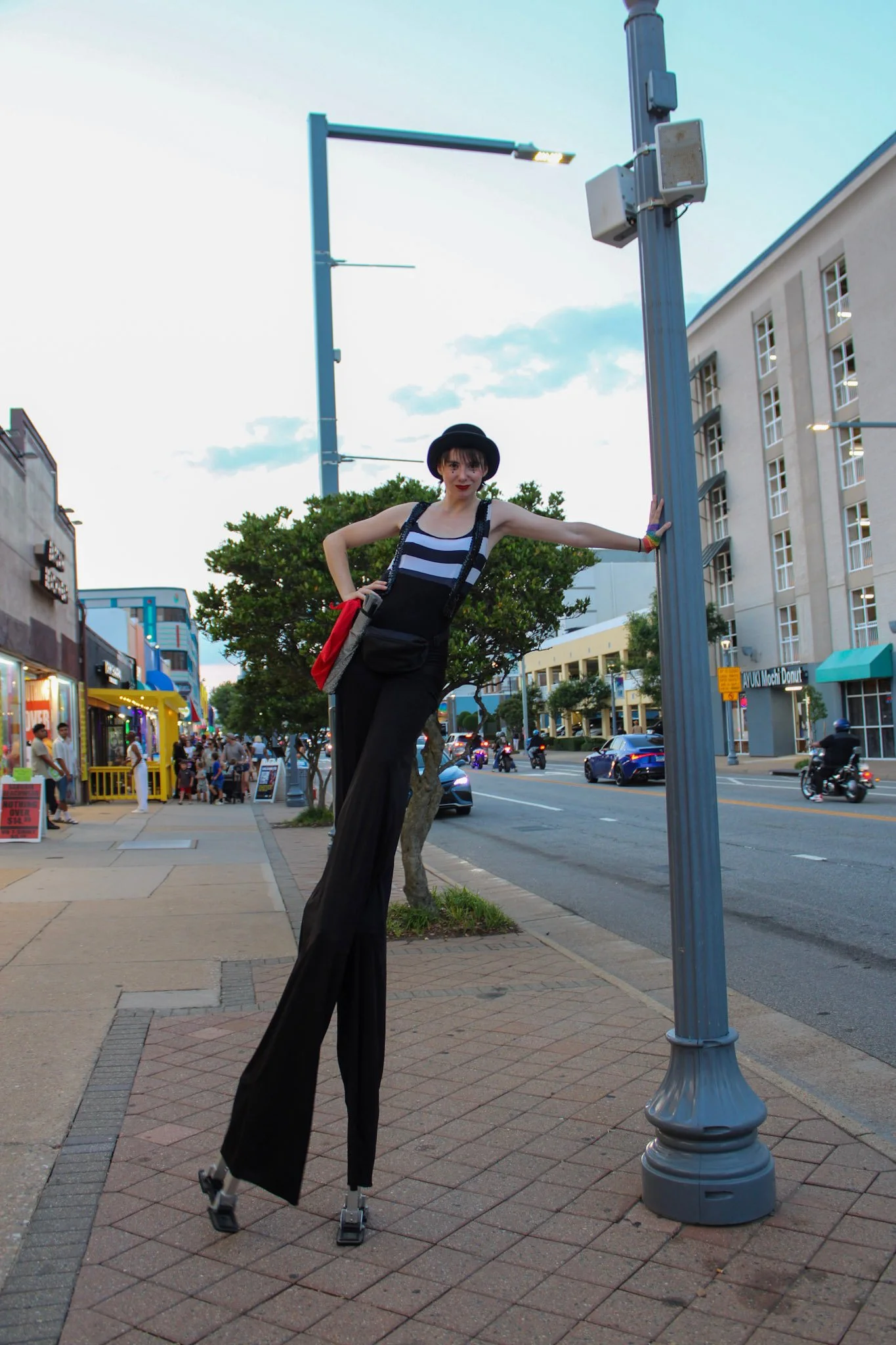 A woman on stilts wearing a black hat and black outfit, standing on a city sidewalk next to a streetlight pole with a blue sky in the background.