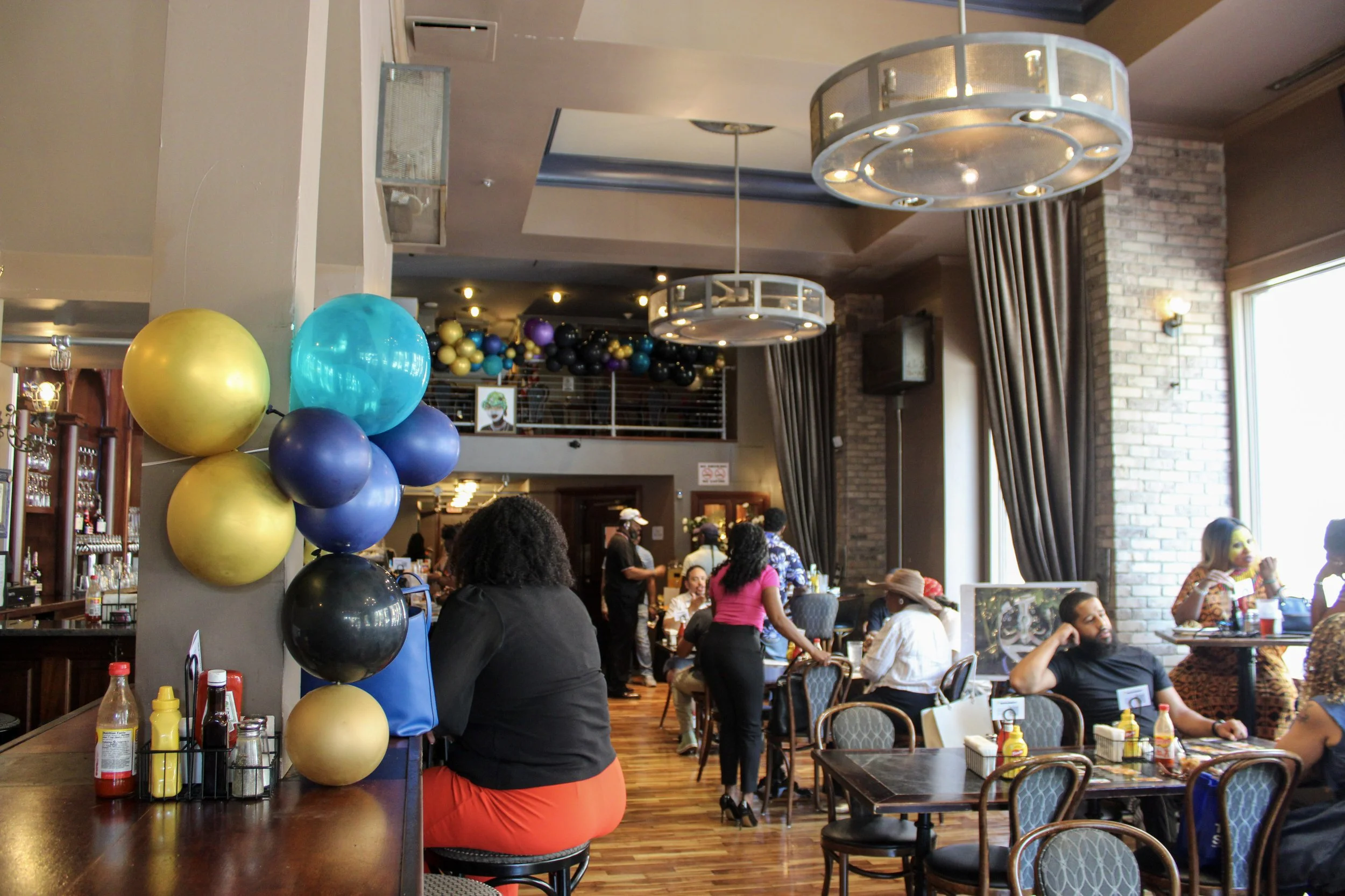 A busy restaurant filled with people, colorful balloons hanging from the ceiling, and a bar area with condiments on the counter.